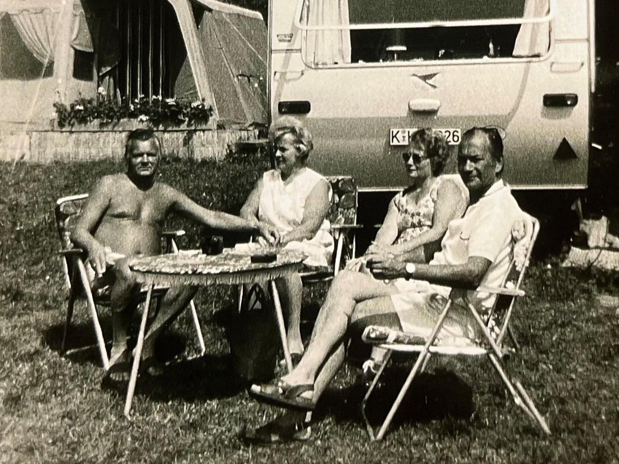 Ein Schwarz-Weiß-Bild zeigt zwei Paar, die auf Campingmöbeln vor einem Wohnwagen sitzen.