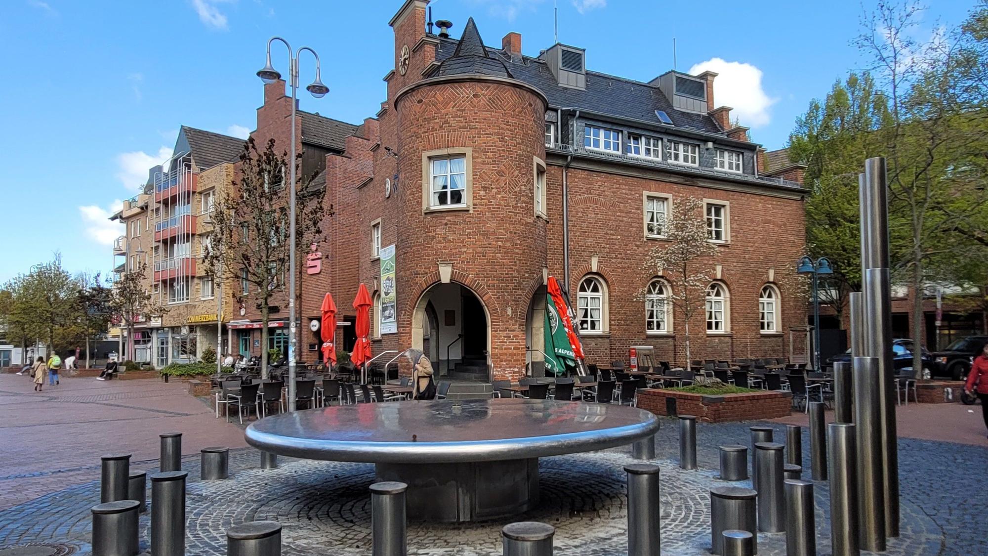 Vorne im Bild ein Brunnen, im Hintergrund ein Backsteingebäude.
