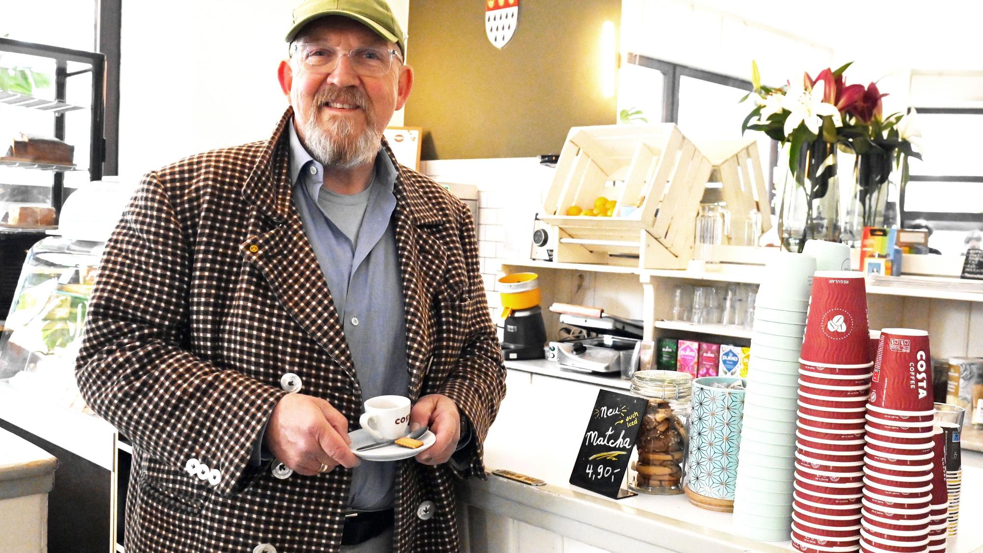 Ein Mann im karierten Sakko steht vor der Theke einer Kaffeebar. In der Hand hält er eine Espresso-Tasse.
