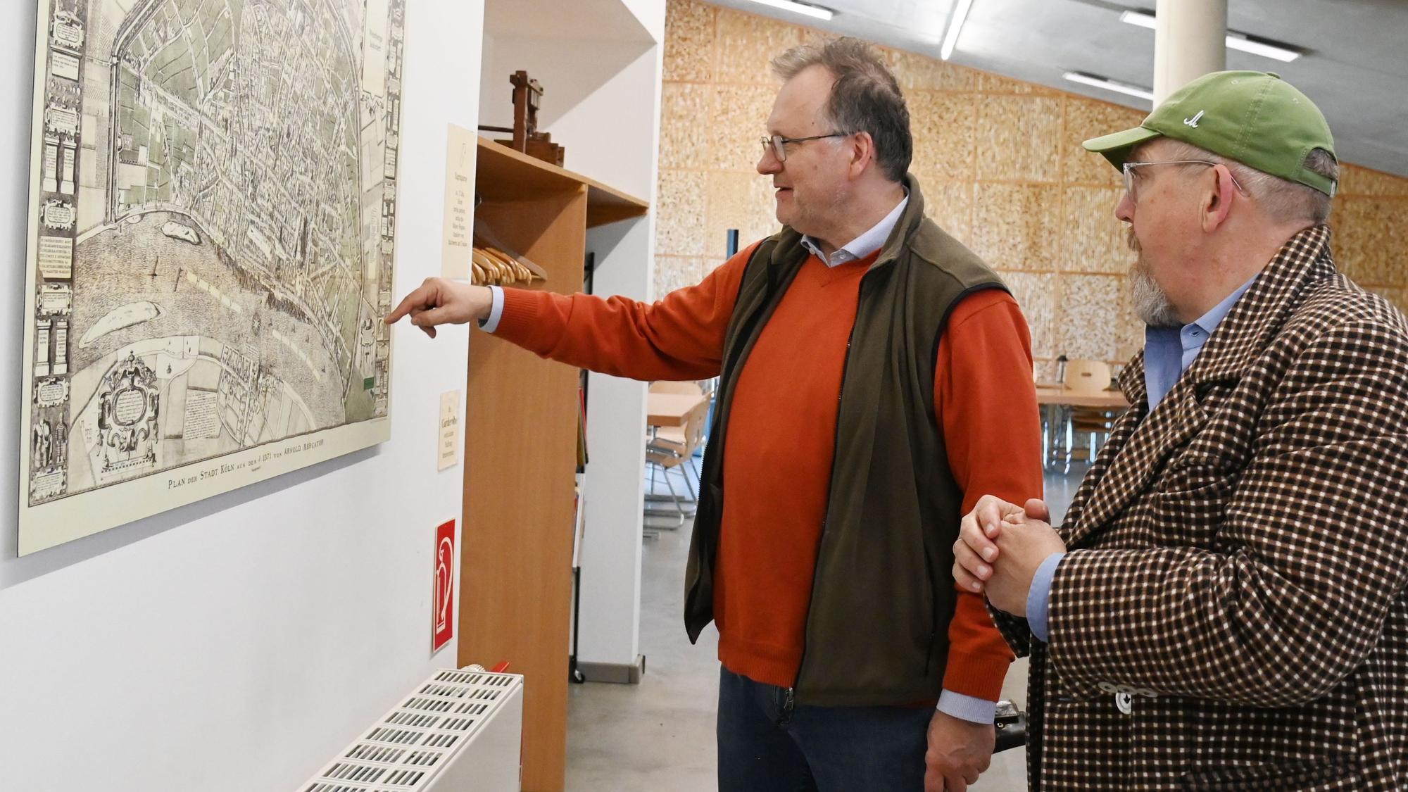 Ein Mann im roten Pullover und Weste zeigt einem Mann mit Bart und Kappe Orte auf einer Karte, die an der Wand hängt.