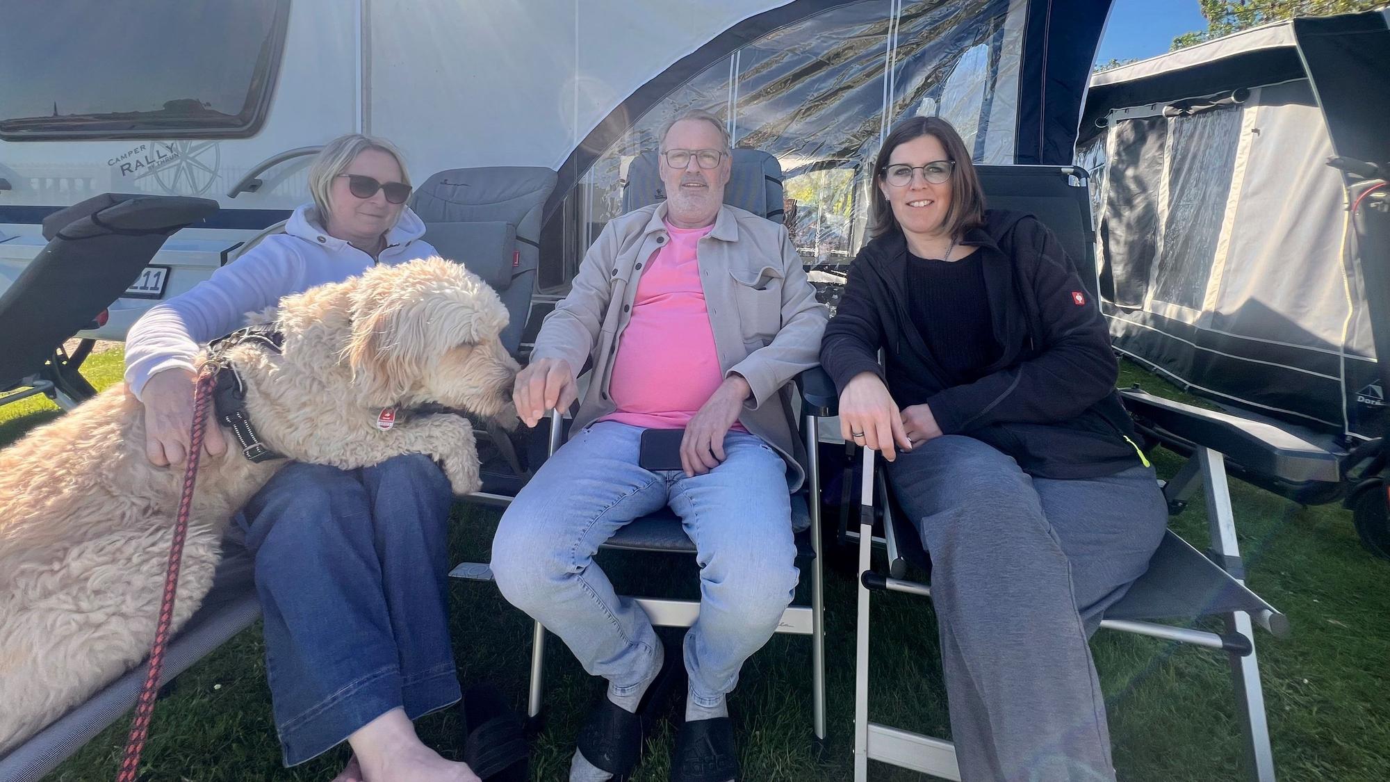 Zwei Frauen, ein Mann und ein großer zotteliger Hund sitzen auf Campingstühlen vor einem Zelt.