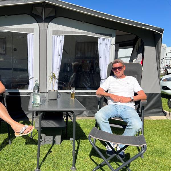 Eine Frau und ein Mann sitzen vor einem Wohnwagen, zwischen sich ein Campingtisch mit Kölsch und Mineralwasser darauf.