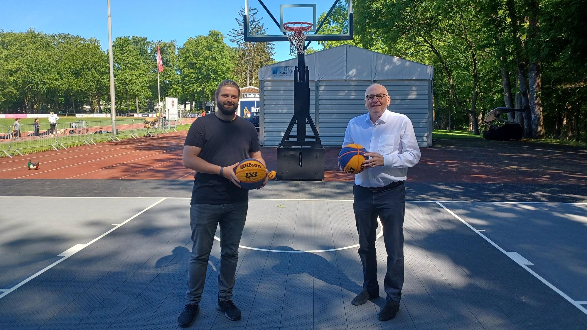 Zwei Männer halten Basketbälle in ihren Händen und stehen auf einem neuen 3x3-Basketballplatz.