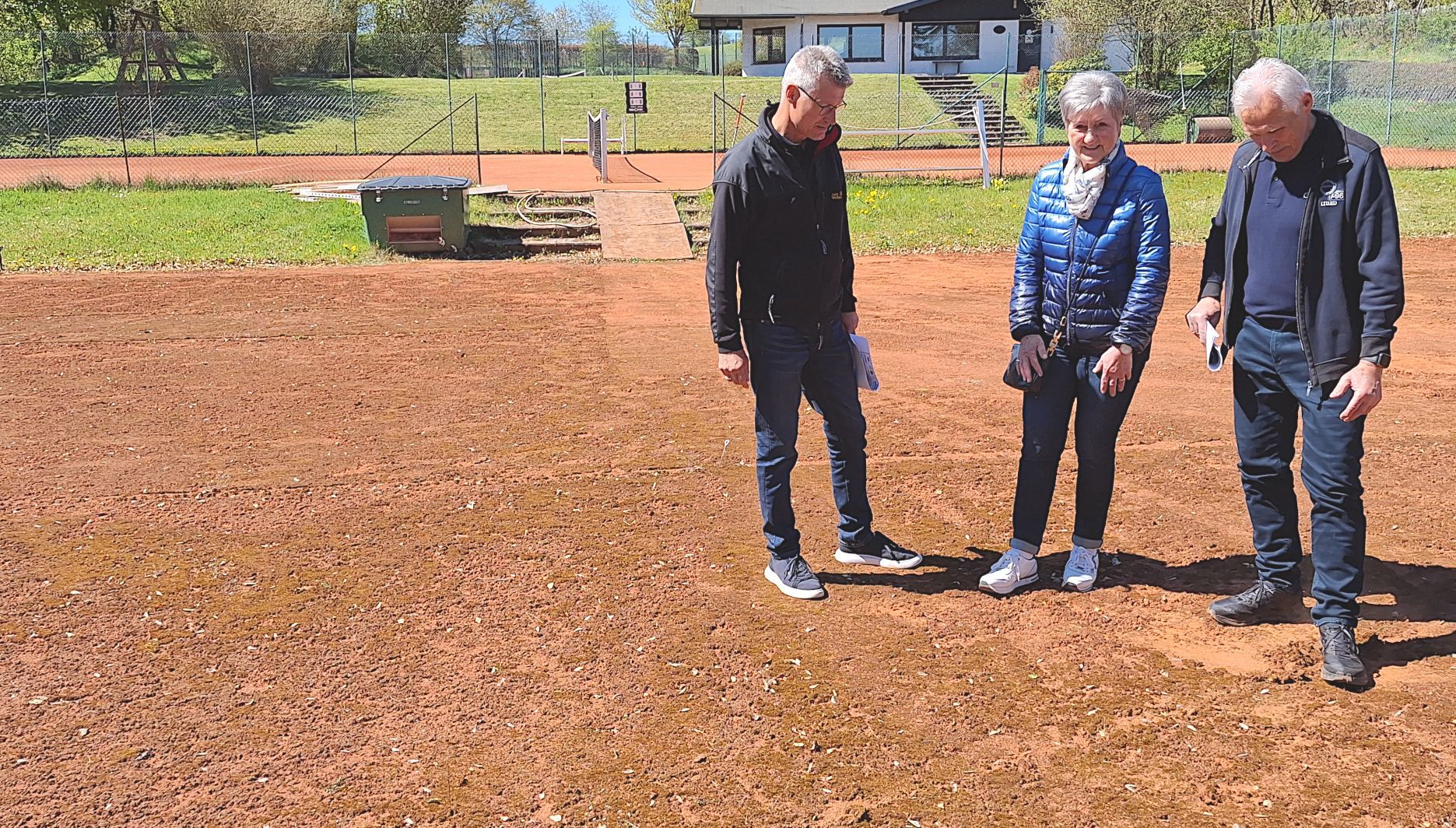 Die drei personen stehen auf einem Tennisplatz und betrachten den rötlichen Belag.