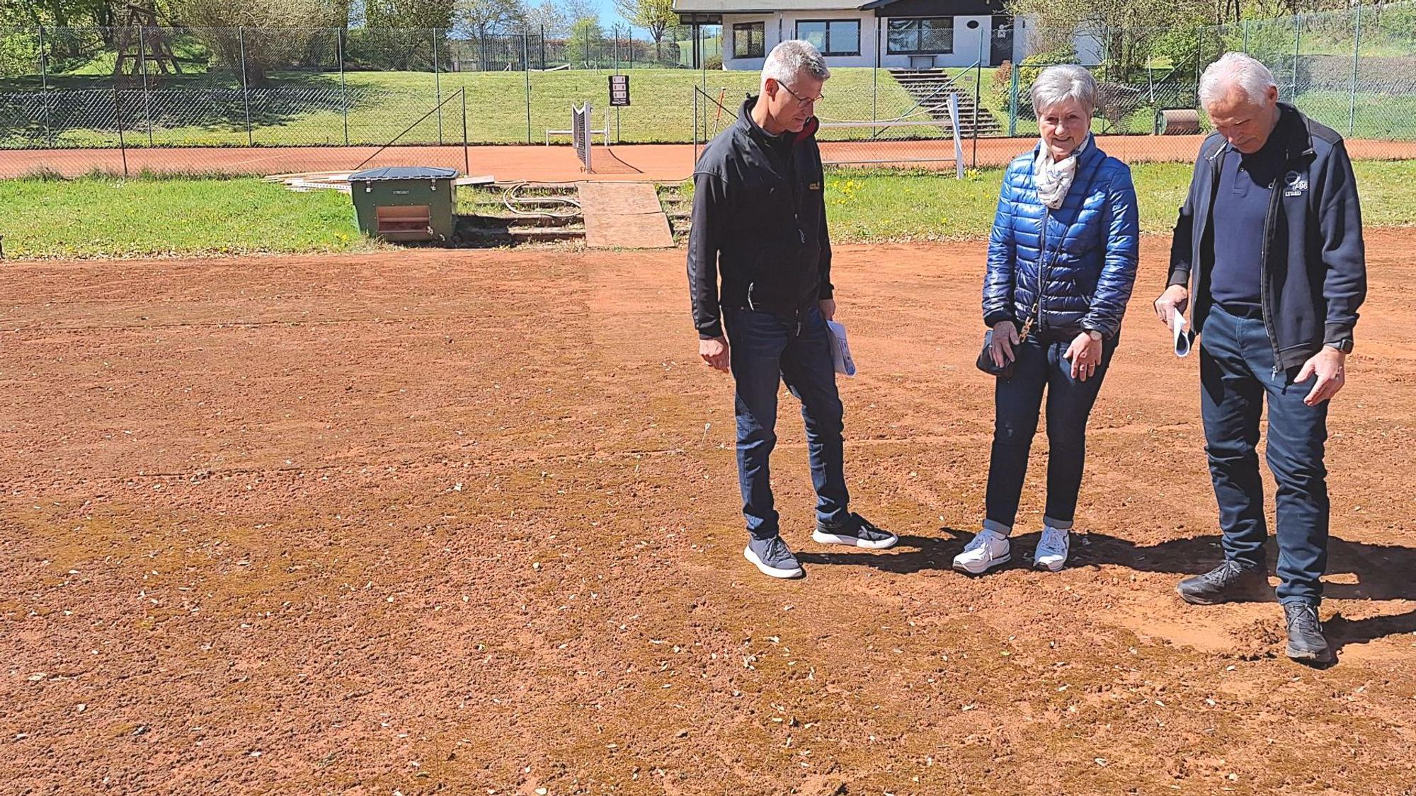 Die drei personen stehen auf einem Tennisplatz und betrachten den rötlichen Belag.