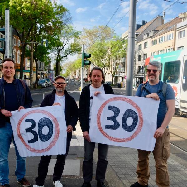 Vier Männer stehen auf einer vielspurigen Straße mit einer Straßenbahn im Hintergrund nebeneinander und halten Plakate mit Tempo-30-Emblem hoch.