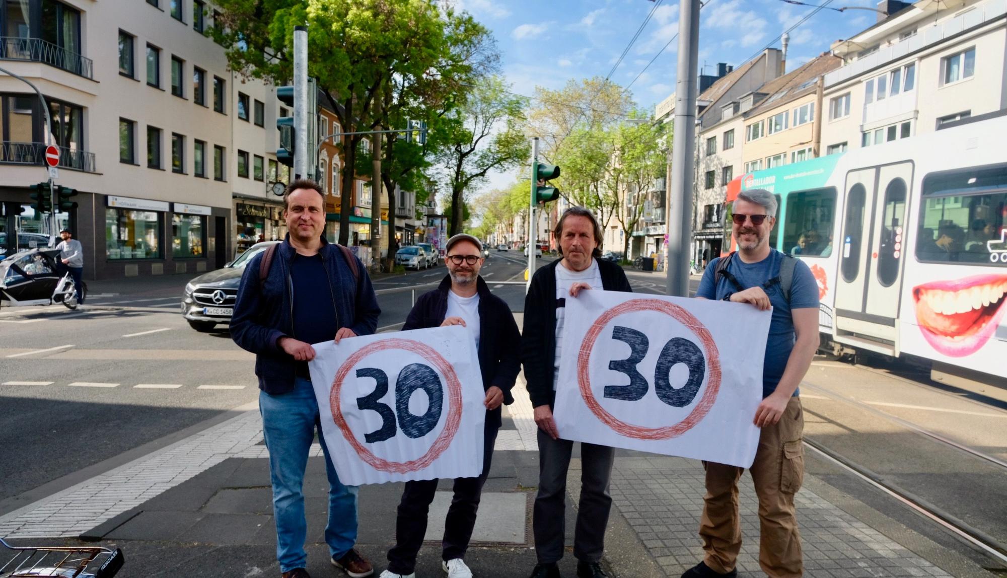 Vier Männer stehen auf einer vielspurigen Straße mit einer Straßenbahn im Hintergrund nebeneinander und halten Plakate mit Tempo-30-Emblem hoch.