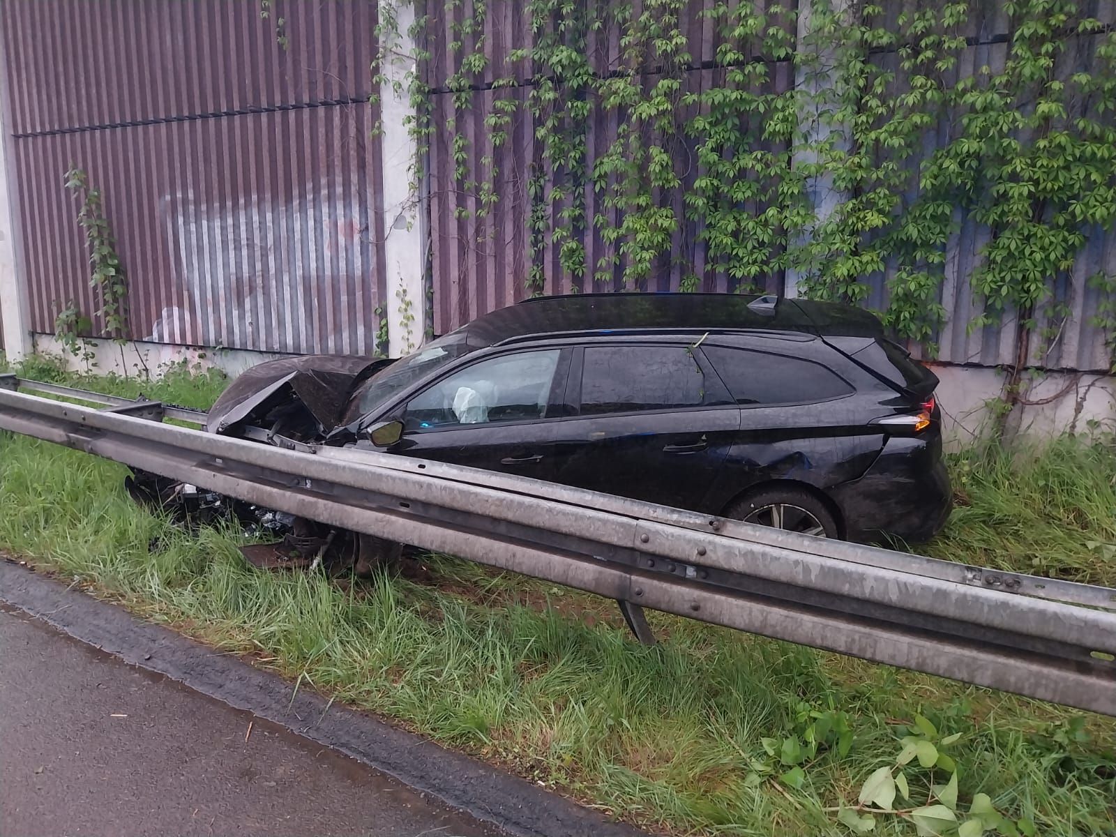 Ein dunkler Kombi steht im Gras hinter der Leitplanke einer Autobahn. Die Motorhaube ist zerbeult.