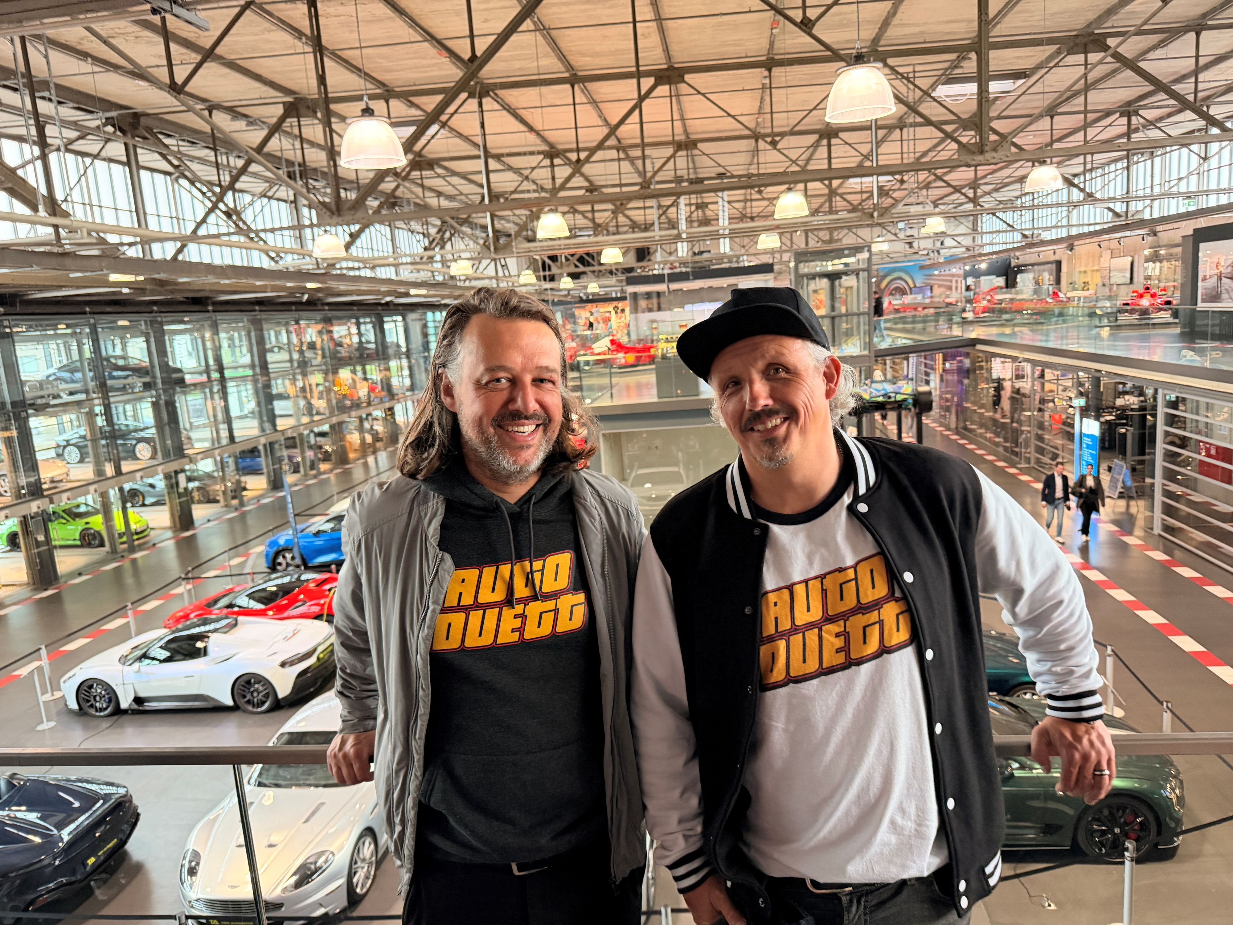 Zwei Männer stehen nebeneinander am Geländer einer Galerie, hinter ihnen sind in einer großen Ausstellungshalle verschiedene Automodelle.