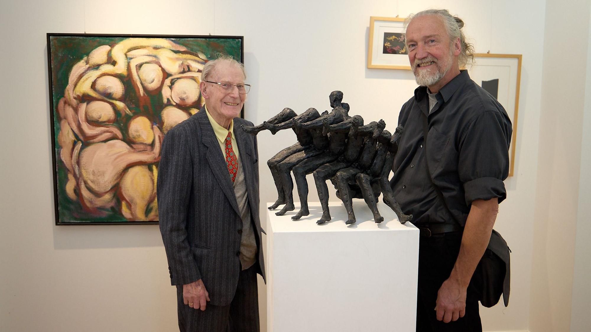 Zwei Männer stehen in einer Galerie neben einer schwarzen Skulpturm, im Hintergrund sind Gemälde zu erkennen.