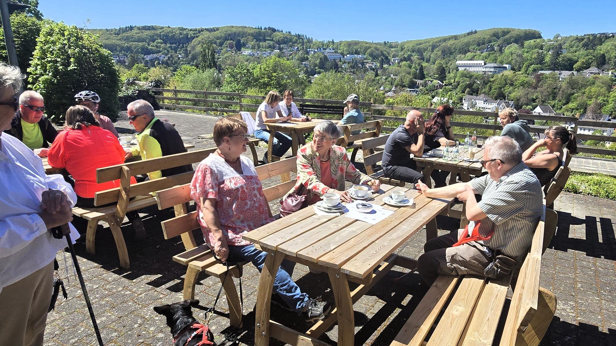 Menschen sitzen auf einer Café-Terrasse in der Sonne und genießen Kaffee, Kuchen und den Ausblick auf Bad Münstereifel.