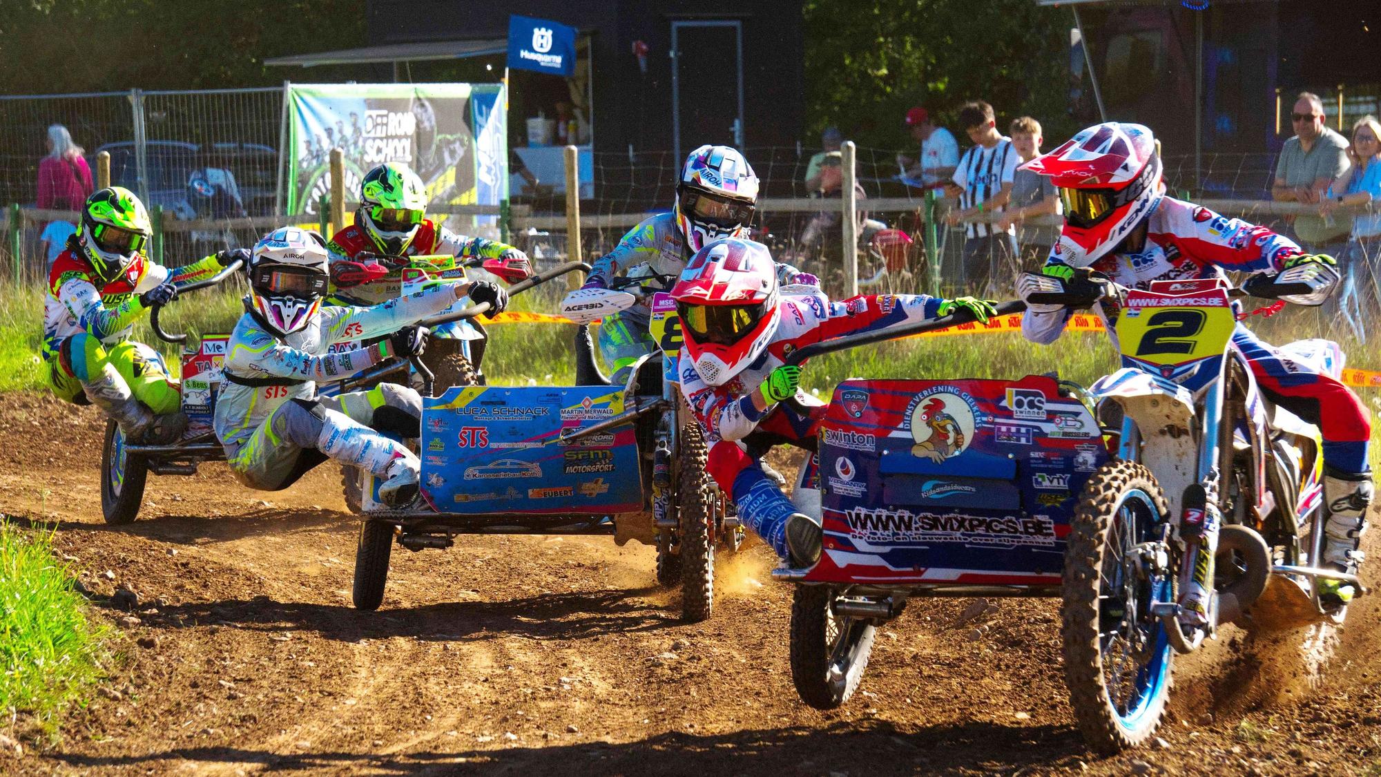Drei Motocross-Maschinen mit Seitenwagen fahren durch eine Kurve, die Beifahrer hängen artistisch am Seitenwagen.