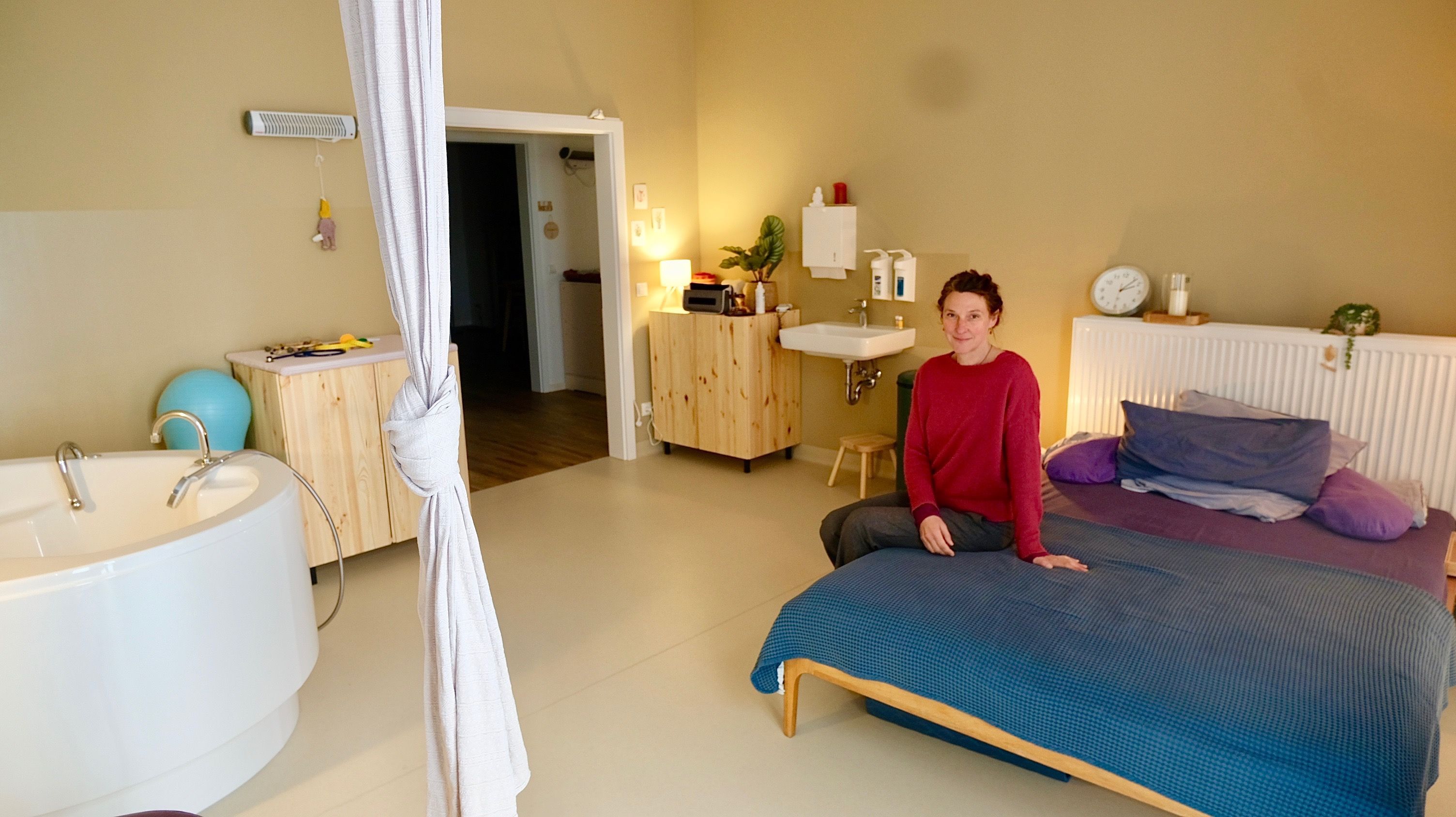 Eine Frau sitzt auf dem großen Bett einem geräumigen Zimmer,  in dem auch eine Badewanne steht.