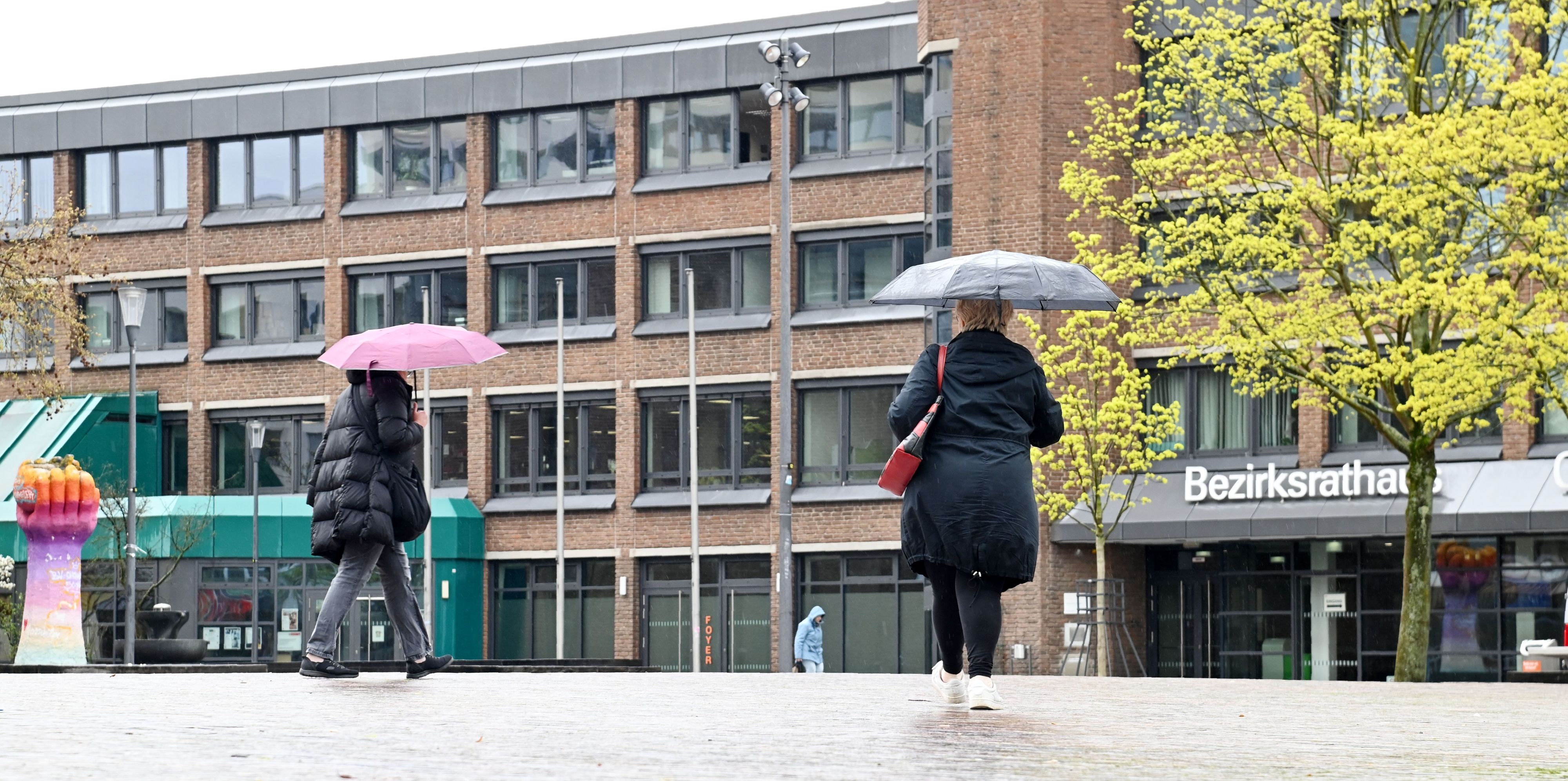 Frauen mit Regenschirmen sind auf einem Platz vor einem mehrstöckigen Gebäude unterwegs.