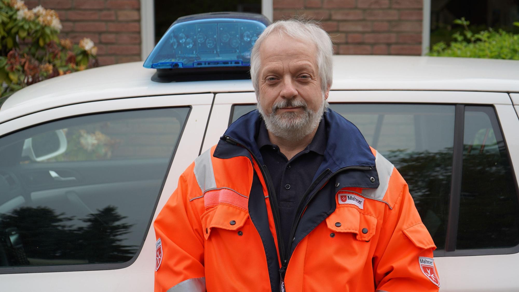 Das Bild zeigt Hornfeck mit einer orangen Jacke vor einem Dienstwagen mit Blaulicht.