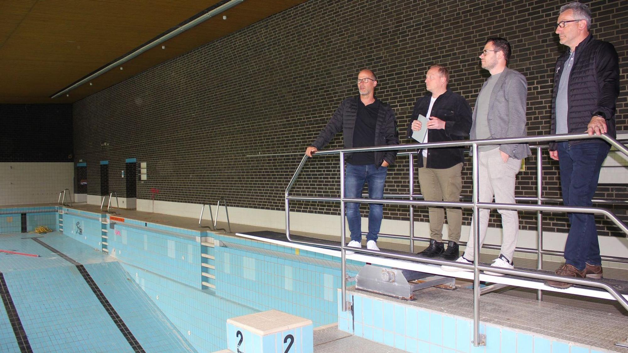 Bei einem Ortstermin im Schwimmbad stehen die vier genannten Männer auf dem Sprungbrett über dem leeren Becken.