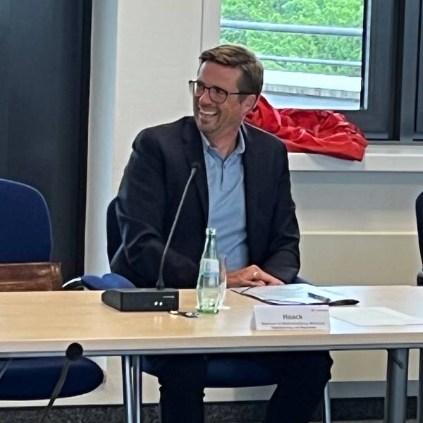 Ein Mann mit Brille und dunklem Jackett sitzt neben einem Mann mit kinnlangem Haar, schwarzem Pullover und buntem Schal.