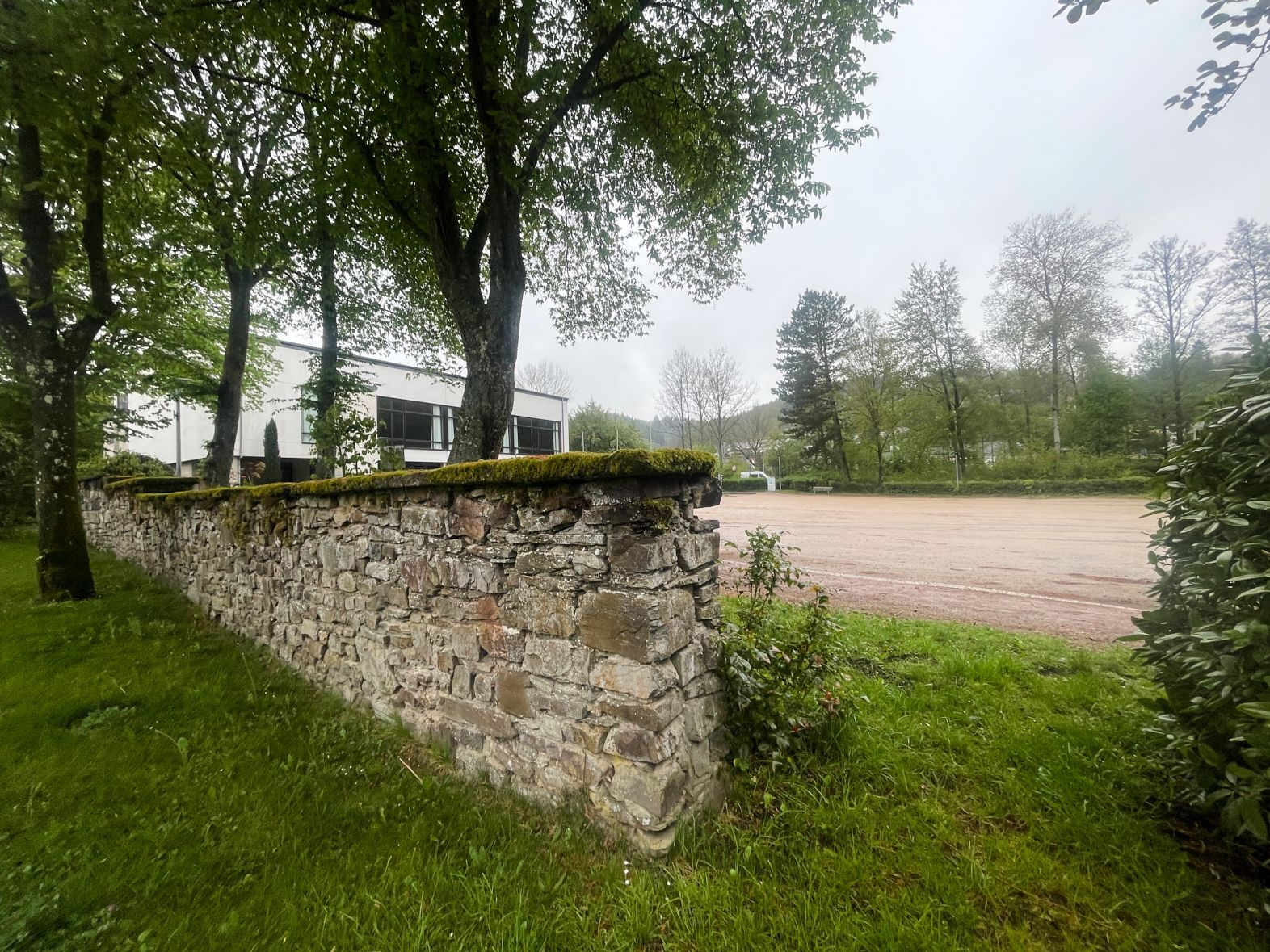 Blick auf das Areal, im Vordergrund eine Bruchsteinmauer. Hinter Bäumen ist die Grenzlandhalle zu erkennen.