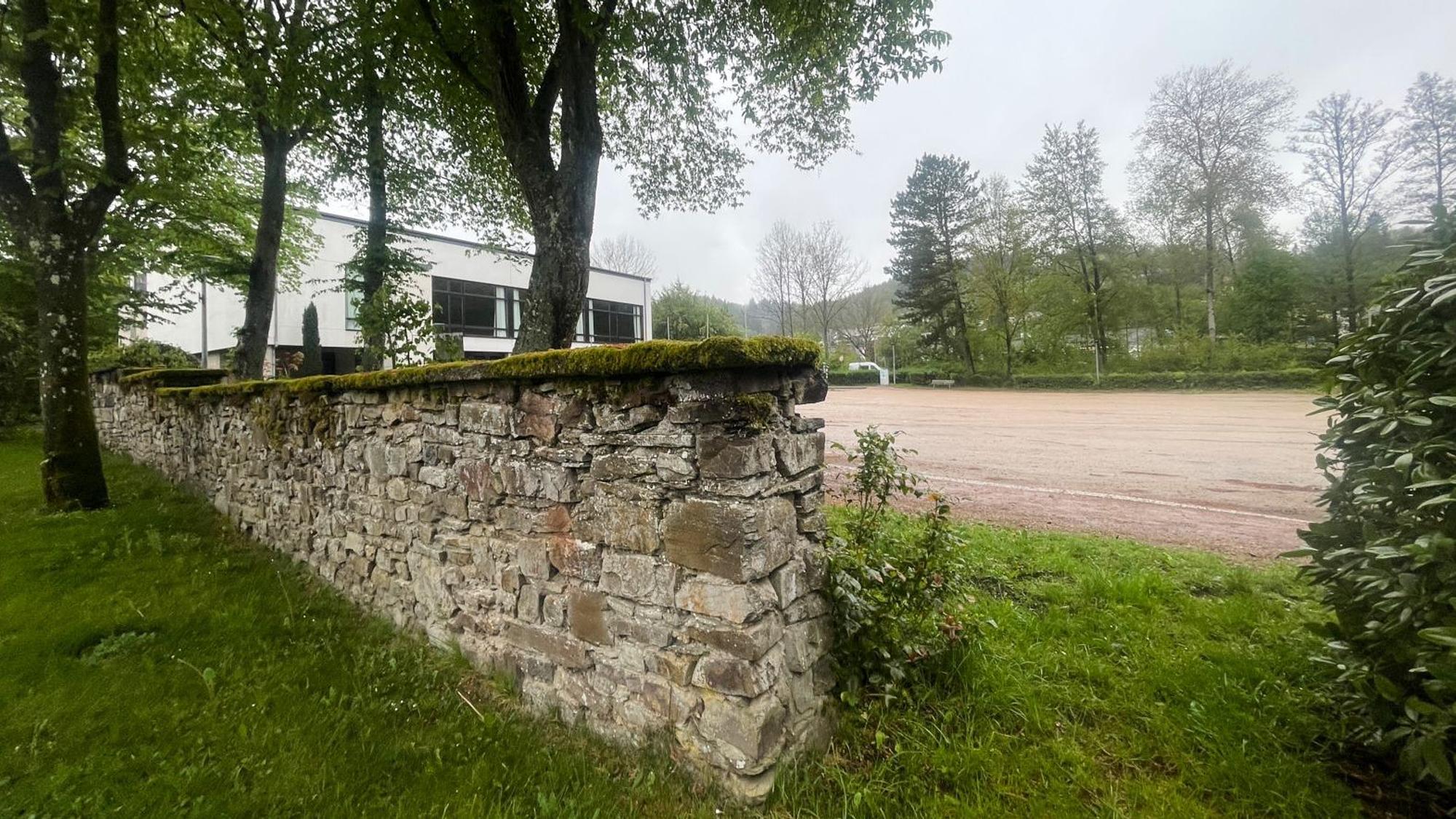 Blick auf das Areal, im Vordergrund eine Bruchsteinmauer. Hinter Bäumen ist die Grenzlandhalle zu erkennen.