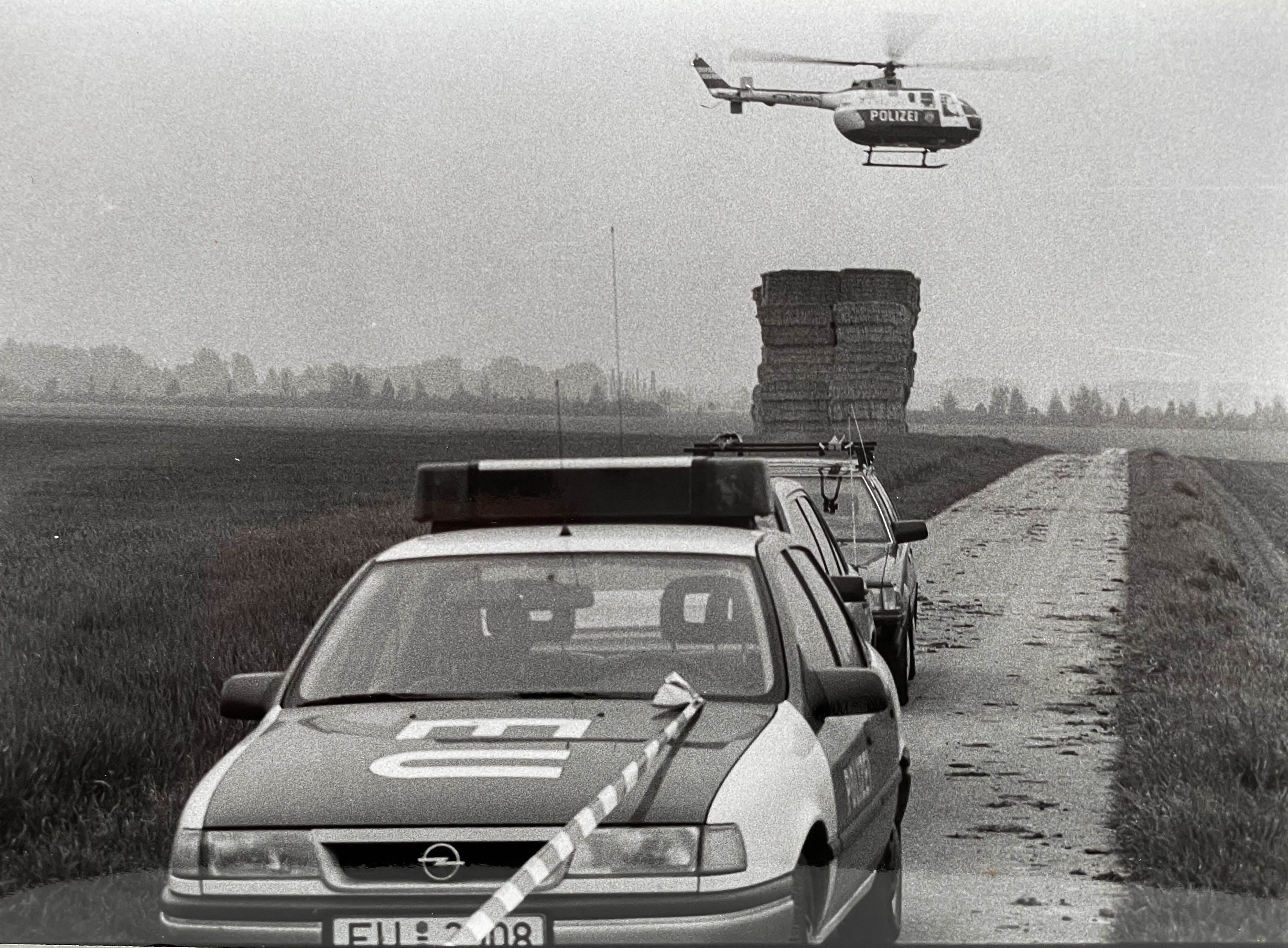 Das Bild zeigt im Vordergrund ein Polizeiauto. Im Hintergrund fliegt ein Polizeihubschrauber.