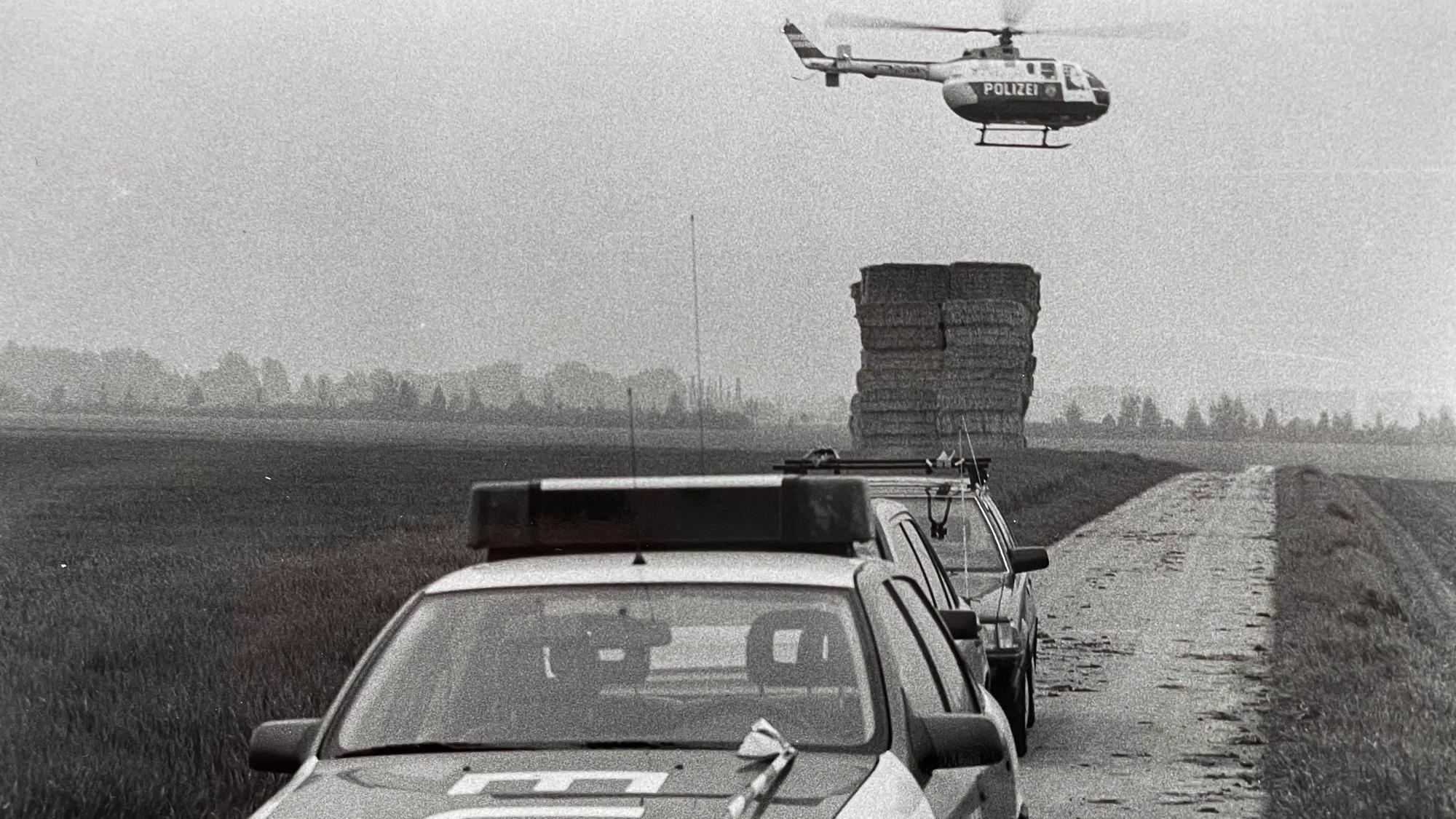 Das Bild zeigt im Vordergrund ein Polizeiauto. Im Hintergrund fliegt ein Polizeihubschrauber.