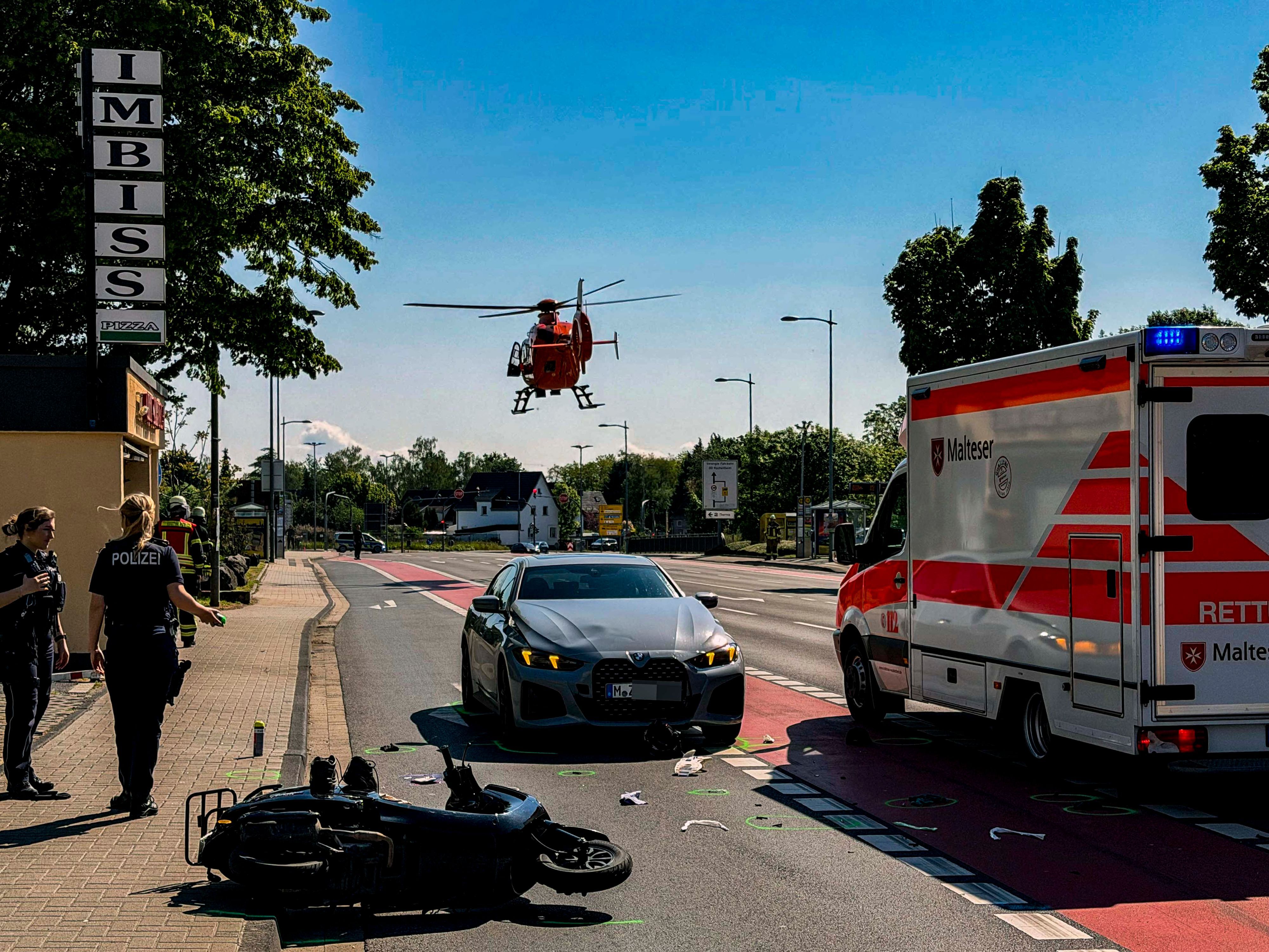 Nach einem Unfall auf der Kölner Straße in Euskirchen liegt ein Motorroller auf der Straße, auf der auch ein BMW und ein Rettungswagen stehen. Ein Rettungshubschrauber setzt gerade zur Landung an.