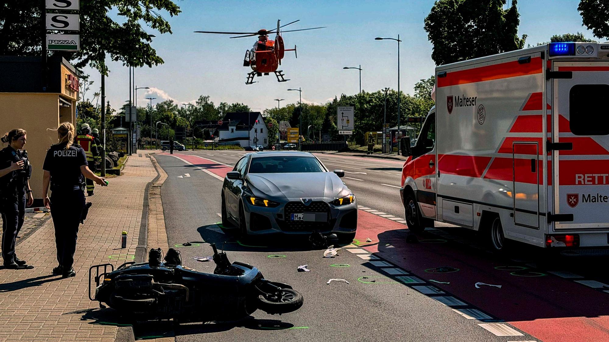 Nach einem Unfall auf der Kölner Straße in Euskirchen liegt ein Motorroller auf der Straße, auf der auch ein BMW und ein Rettungswagen stehen. Ein Rettungshubschrauber setzt gerade zur Landung an.