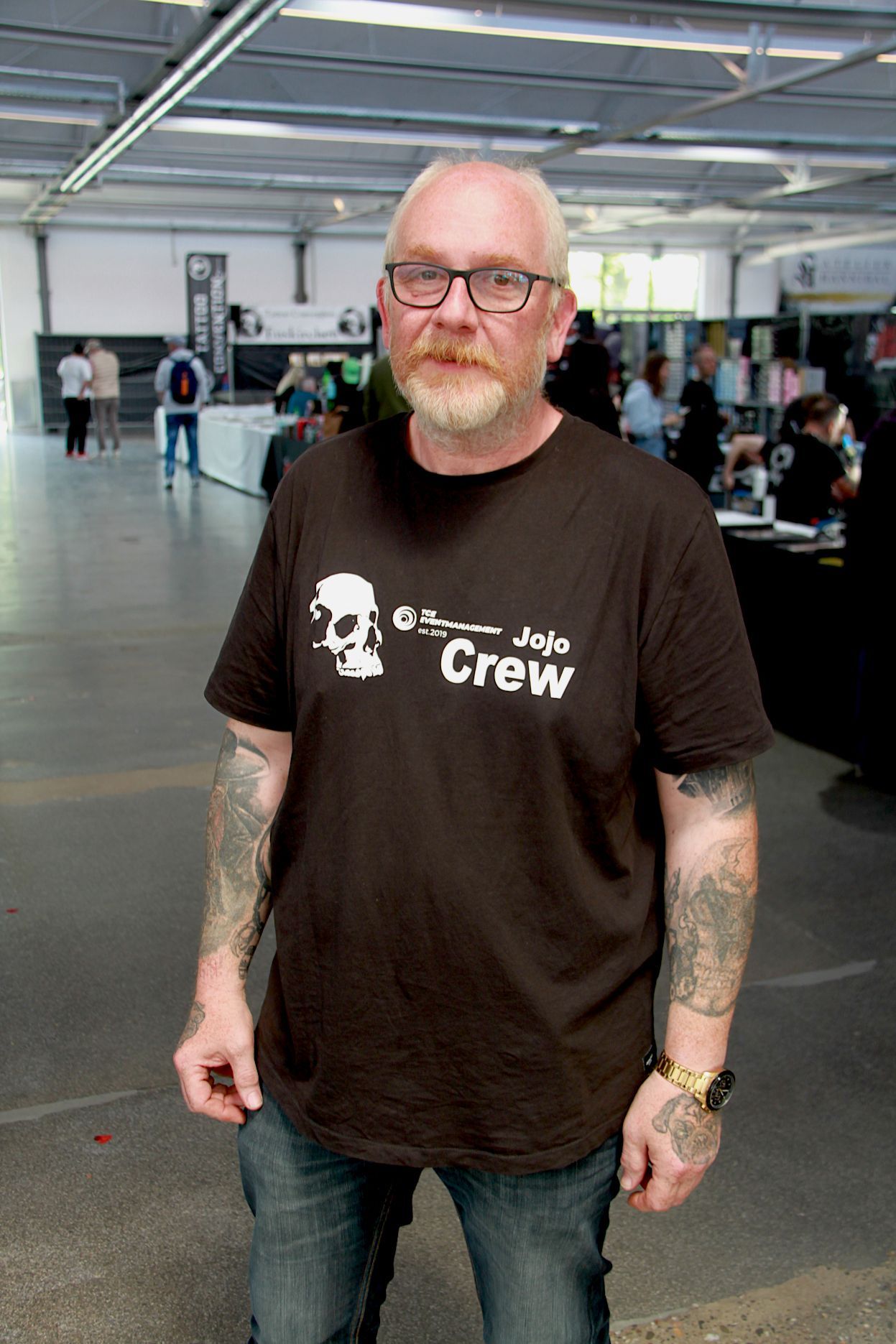 Jürgen Kastenholz tägt Halbglatze, Brille, Bart und ein schwarzes T-Shirt mit weißem Totenkopf-Aufdruck.Seite Arme sind tätowiert.