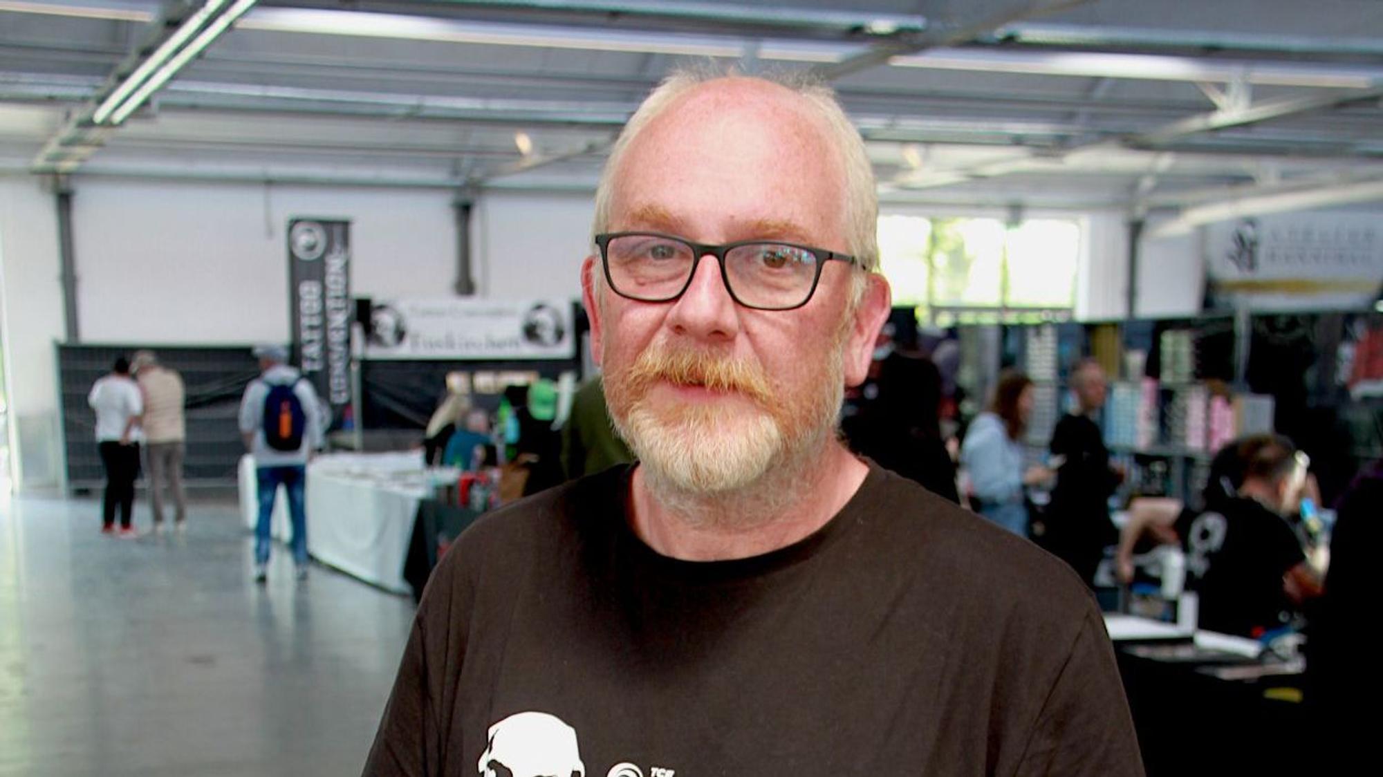 Jürgen Kastenholz tägt Halbglatze, Brille, Bart und ein schwarzes T-Shirt mit weißem Totenkopf-Aufdruck.Seite Arme sind tätowiert.