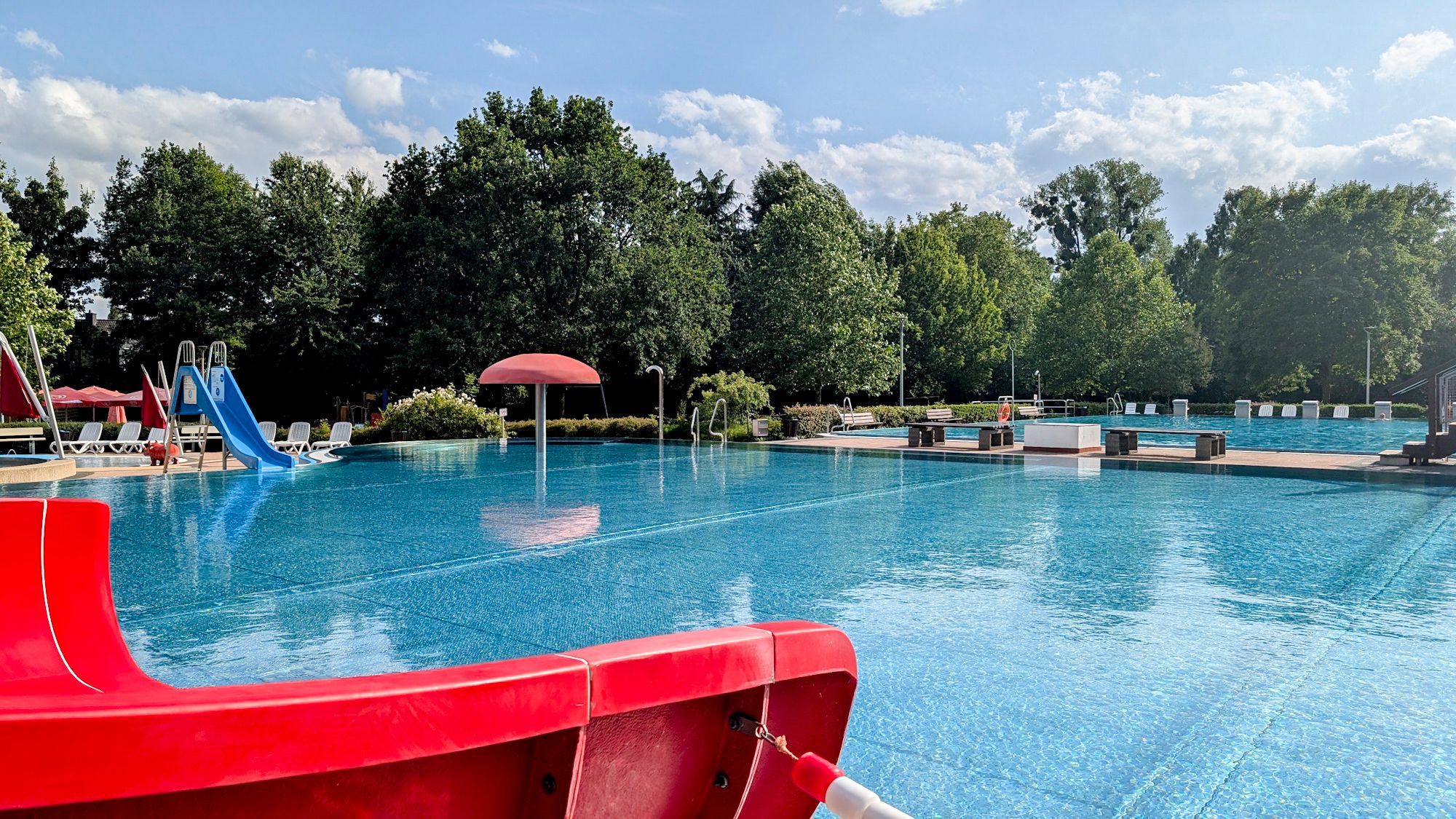 Das Bild zeigt das Schwimmbad, eine blaue Rutsche und einen Pilz im Wasser.