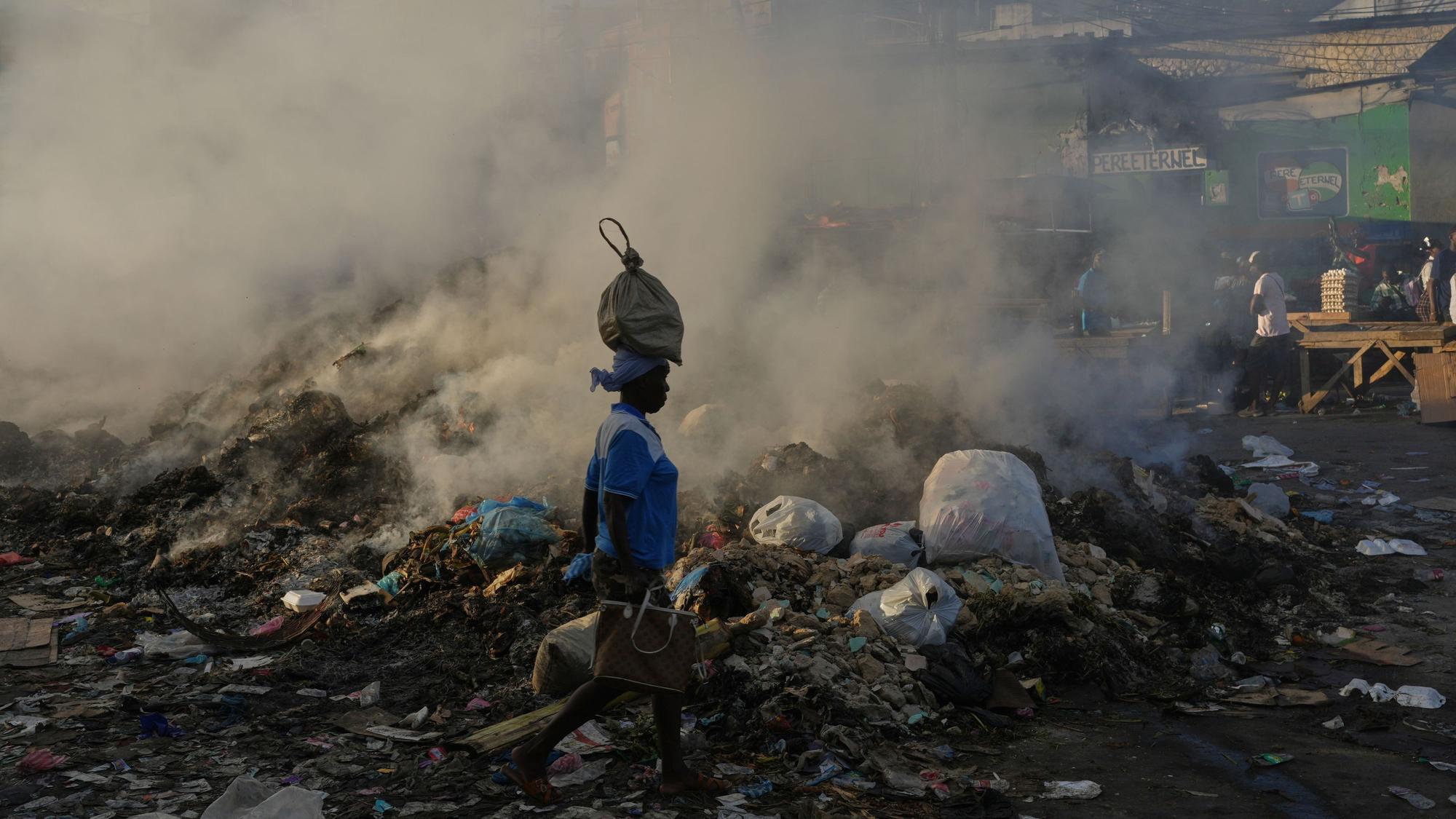 Eine Frau, die eine Tasche auf dem Kopf trägt, geht im Stadtteil Petion-Ville von Port-au-Prince an schwelendem Müll vorbei.