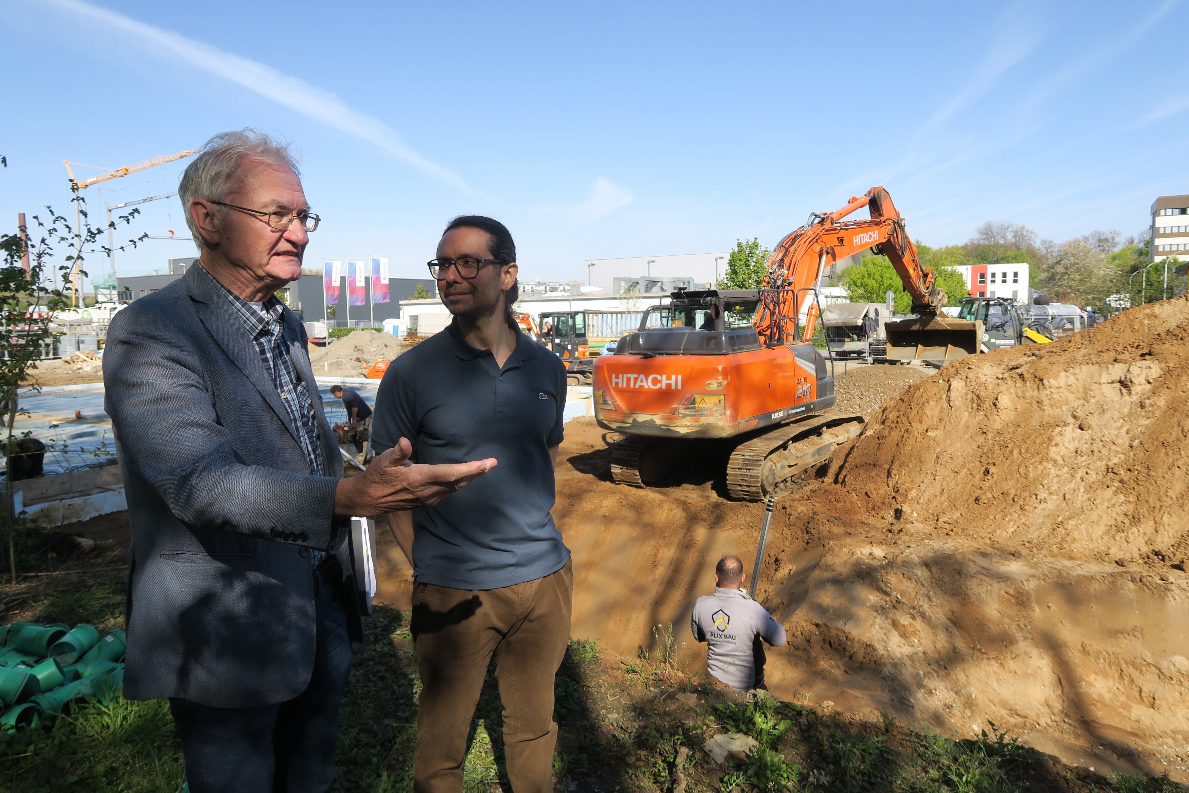 Zwei Männer stehen vor einer großen Baugrube, in der ein Bagger manövriert.