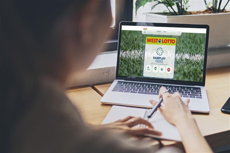 Eine Frau sitzt an einem Tisch und schaut auf ihren Laptop. Der Laptop zeigt die Internetseite des WestLotto Fairplay-Pokals.