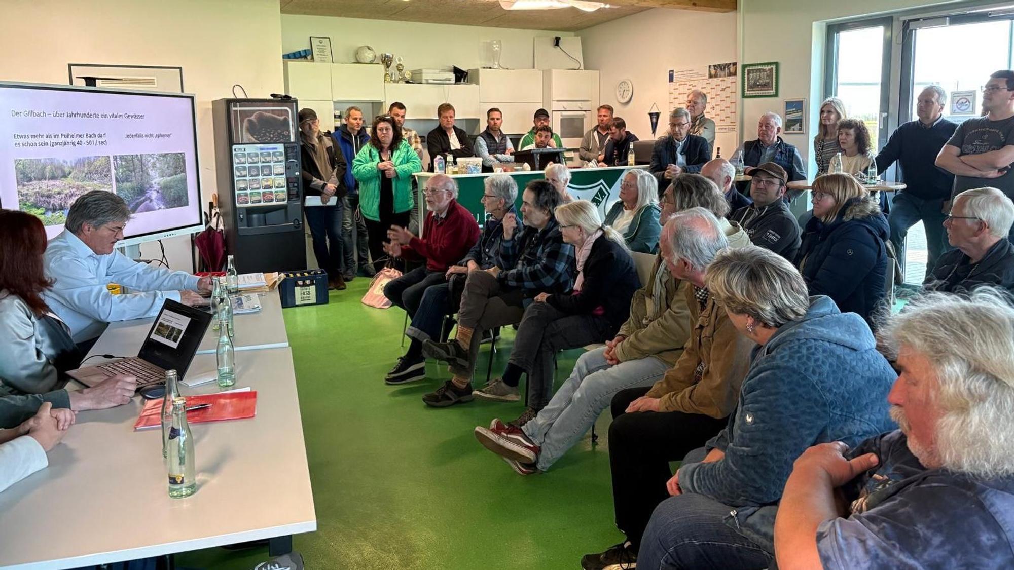 Das Bild zeigt eine Gruppe Menschen in einem Raum. Links im Bild zeigt eine Leinwand Bilder des Gillbach, rechts besetzte Stuhlreihen und einige stehende Gäste.