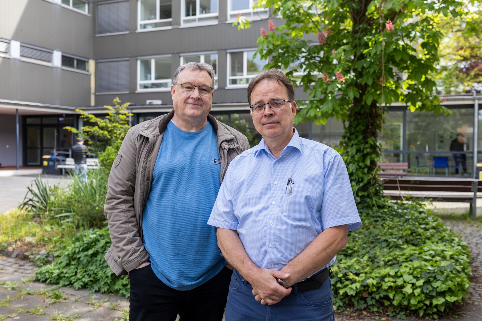 Zwei Männer schauen in die Kamera. Hinter ihnen ein Schulhof.
