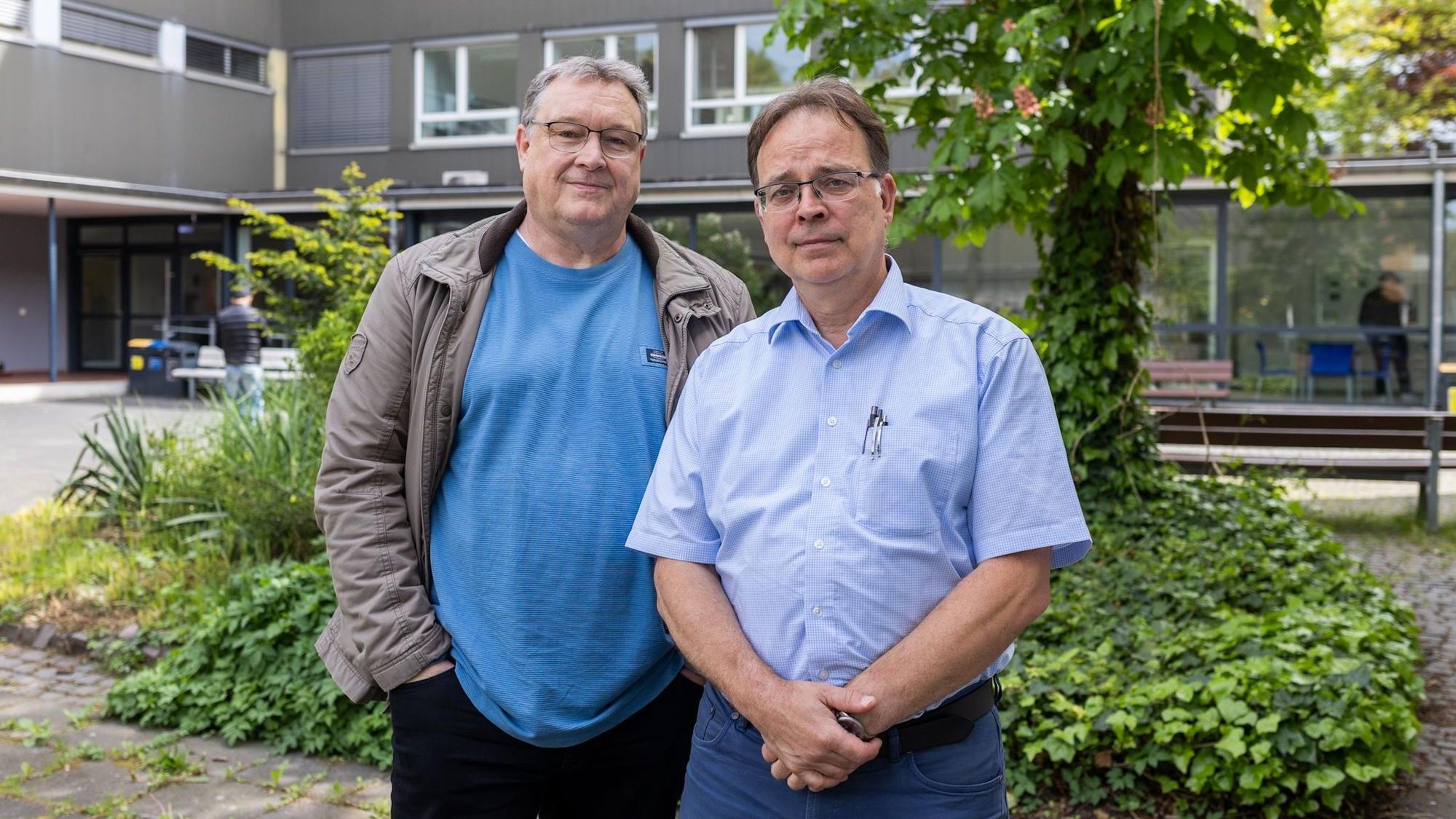 Zwei Männer schauen in die Kamera. Hinter ihnen ein Schulhof.