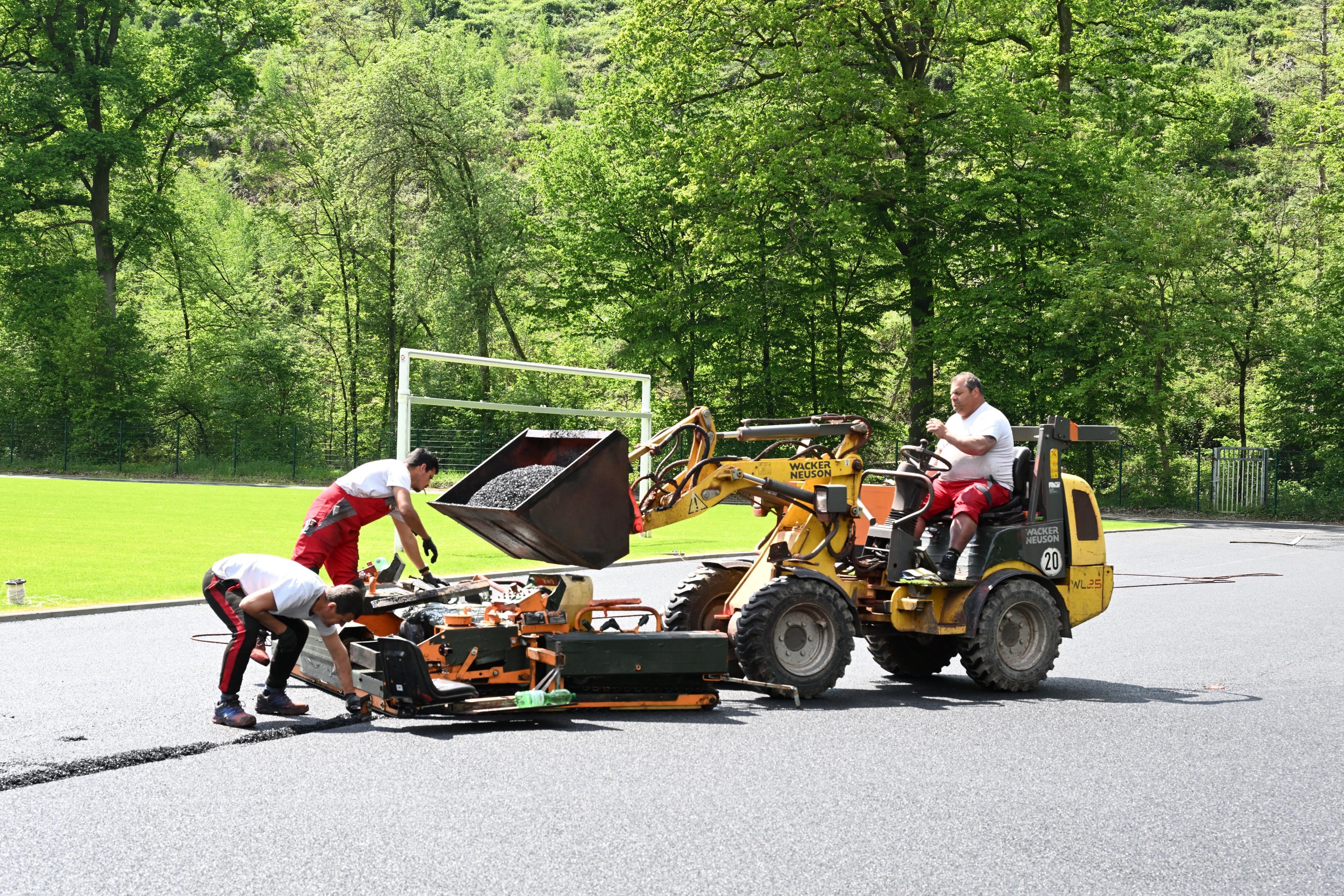 Ein Mann sitzt auf einem Schaufelbagger und lädt dunkles Granulat in eine Mschine, mit der zwei andere Männer das Material auf den Laufbahnen eines Sportplatzes verteilen.