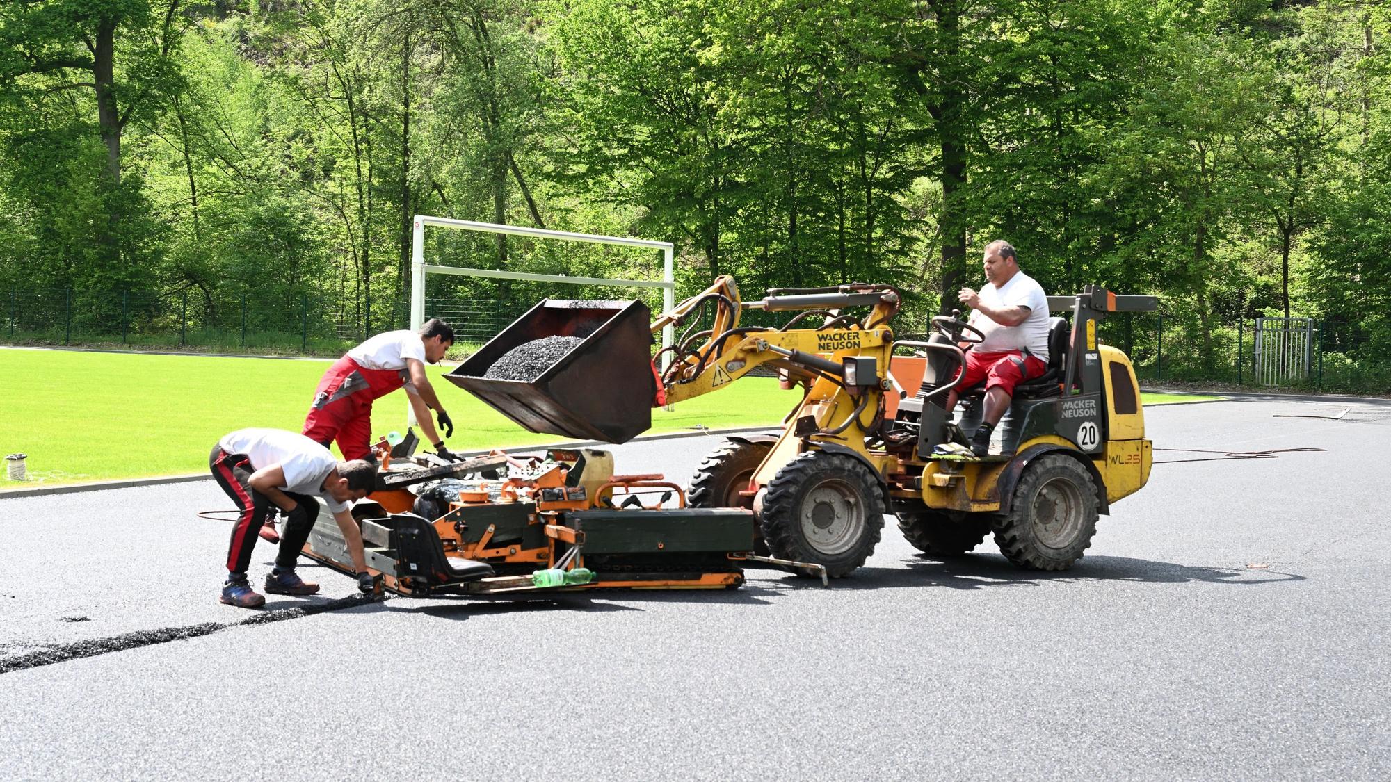 Ein Mann sitzt auf einem Schaufelbagger und lädt dunkles Granulat in eine Mschine, mit der zwei andere Männer das Material auf den Laufbahnen eines Sportplatzes verteilen.