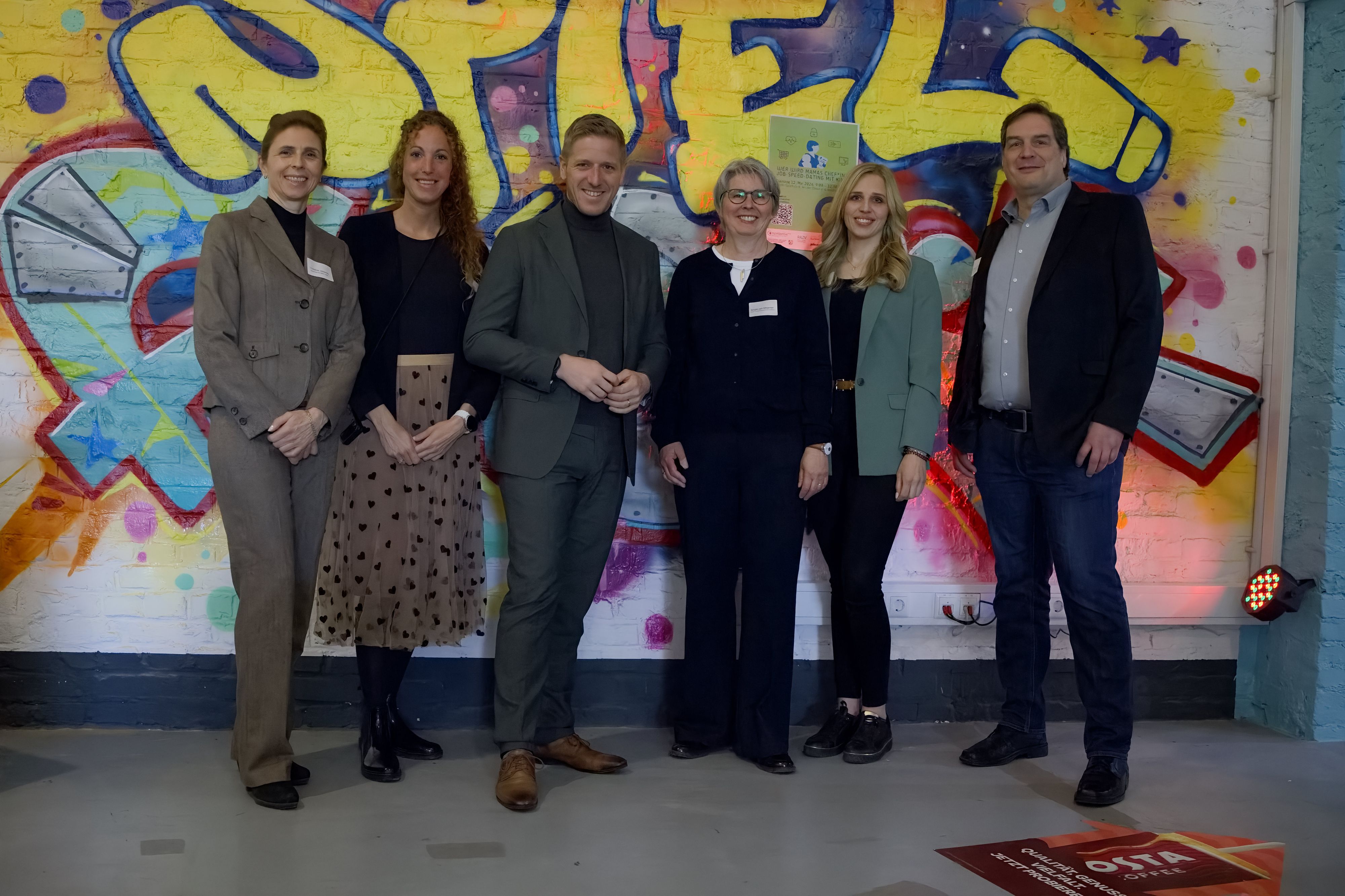 Vier Frauen und zwei Männer stehen in der Zikkurat in Firmenich vor einer Wand, auf der ein Graffiti zu sehen ist.