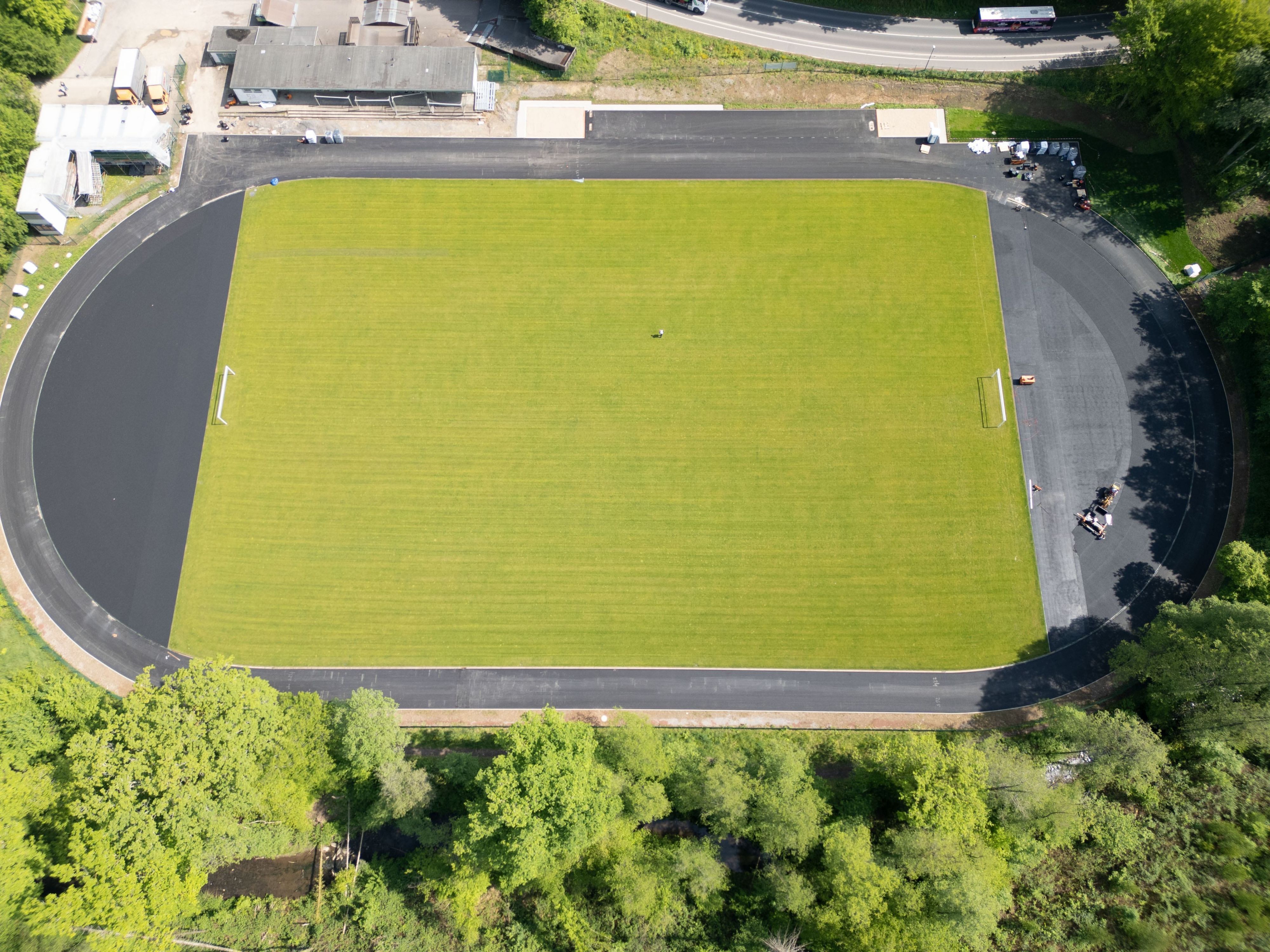Ein im Bau befindlicher Sportplatz aus der Vorgelperspektive. In der Mitte liegt schon grüner Rollrasen, die Laufbahnen werden gerade mit ein einer grau-schwarzen Kunststoffschicht belegt.