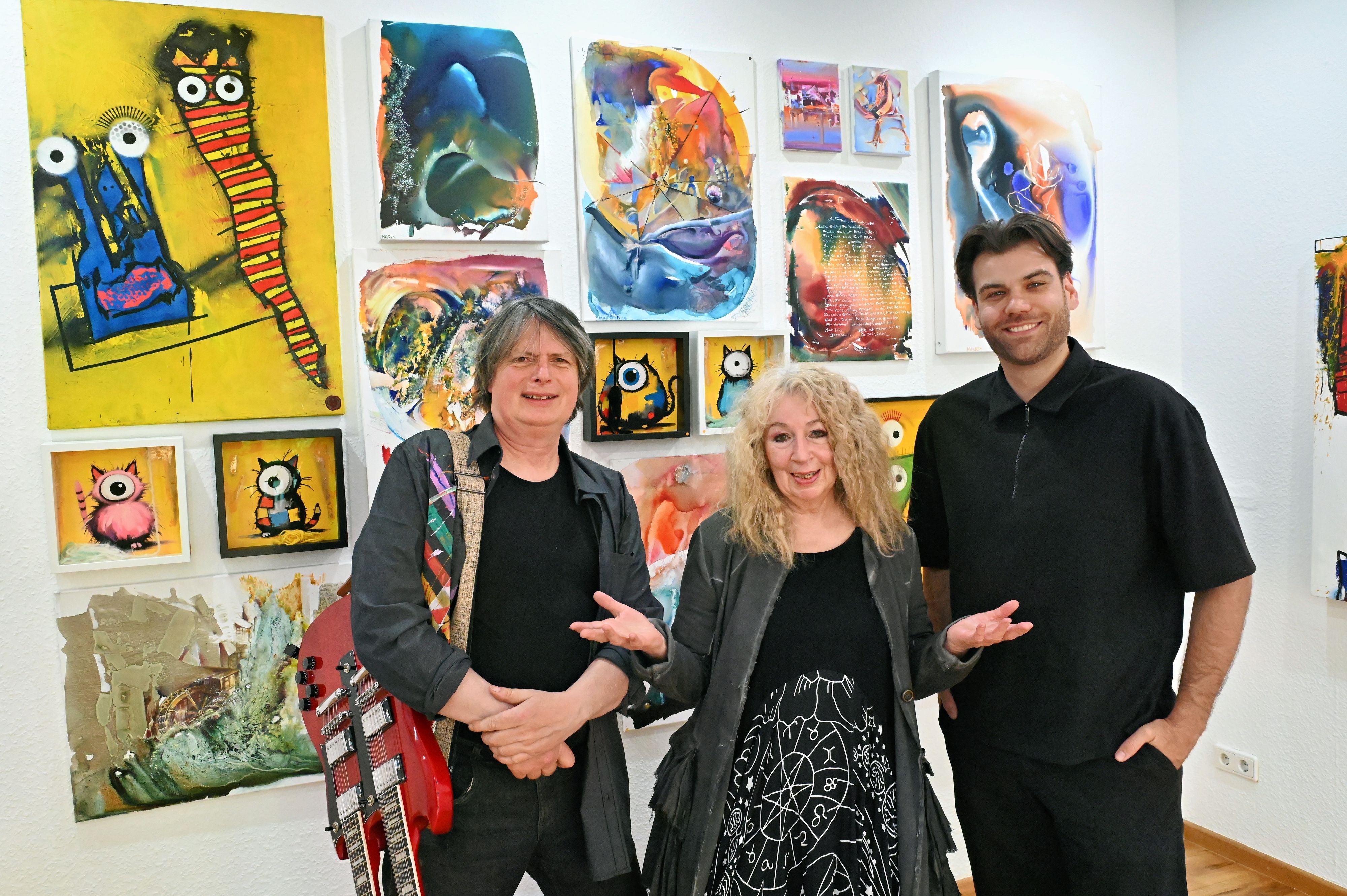 Zwei Männer und eine Frau stehen in der Galerie Roy in Zülpich vor einer Wand, an der zahlreiche Kunstwerke hängen.