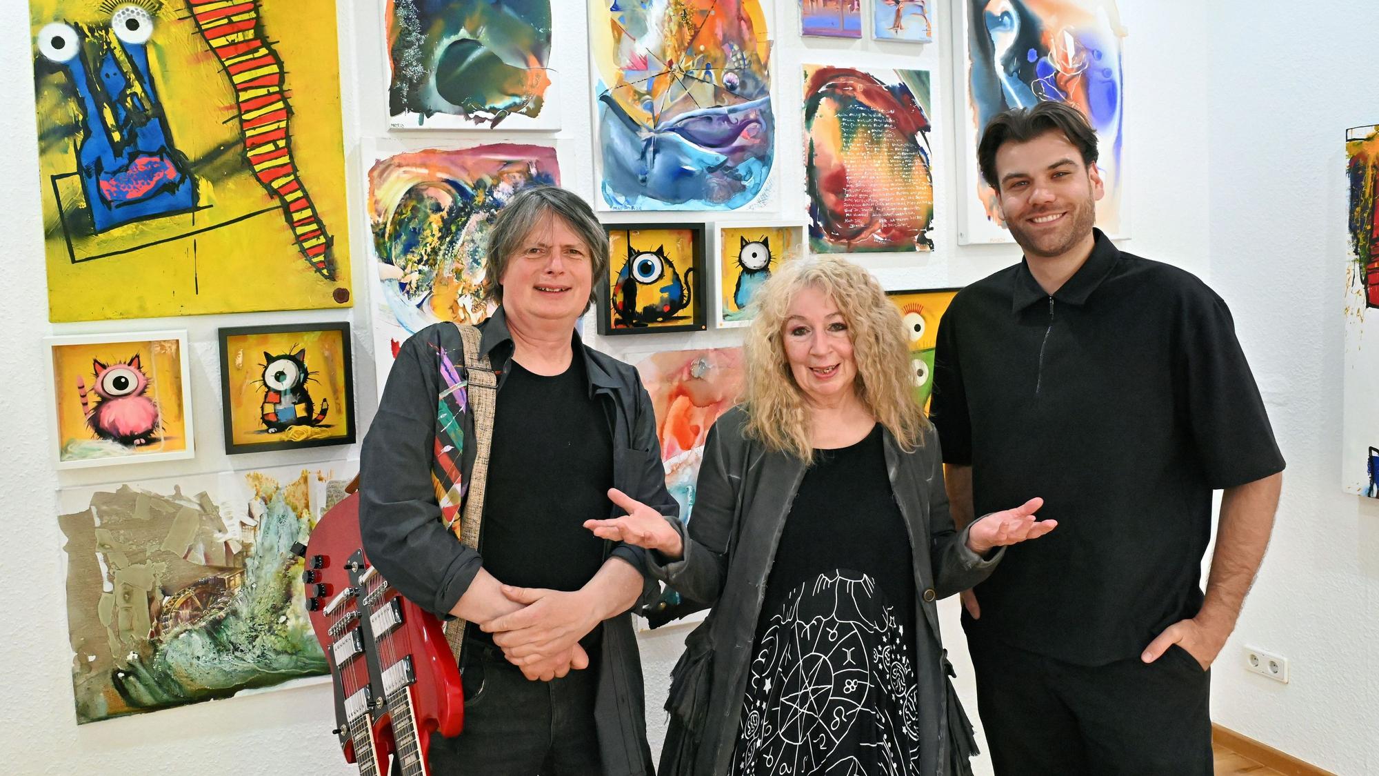 Zwei Männer und eine Frau stehen in der Galerie Roy in Zülpich vor einer Wand, an der zahlreiche Kunstwerke hängen.