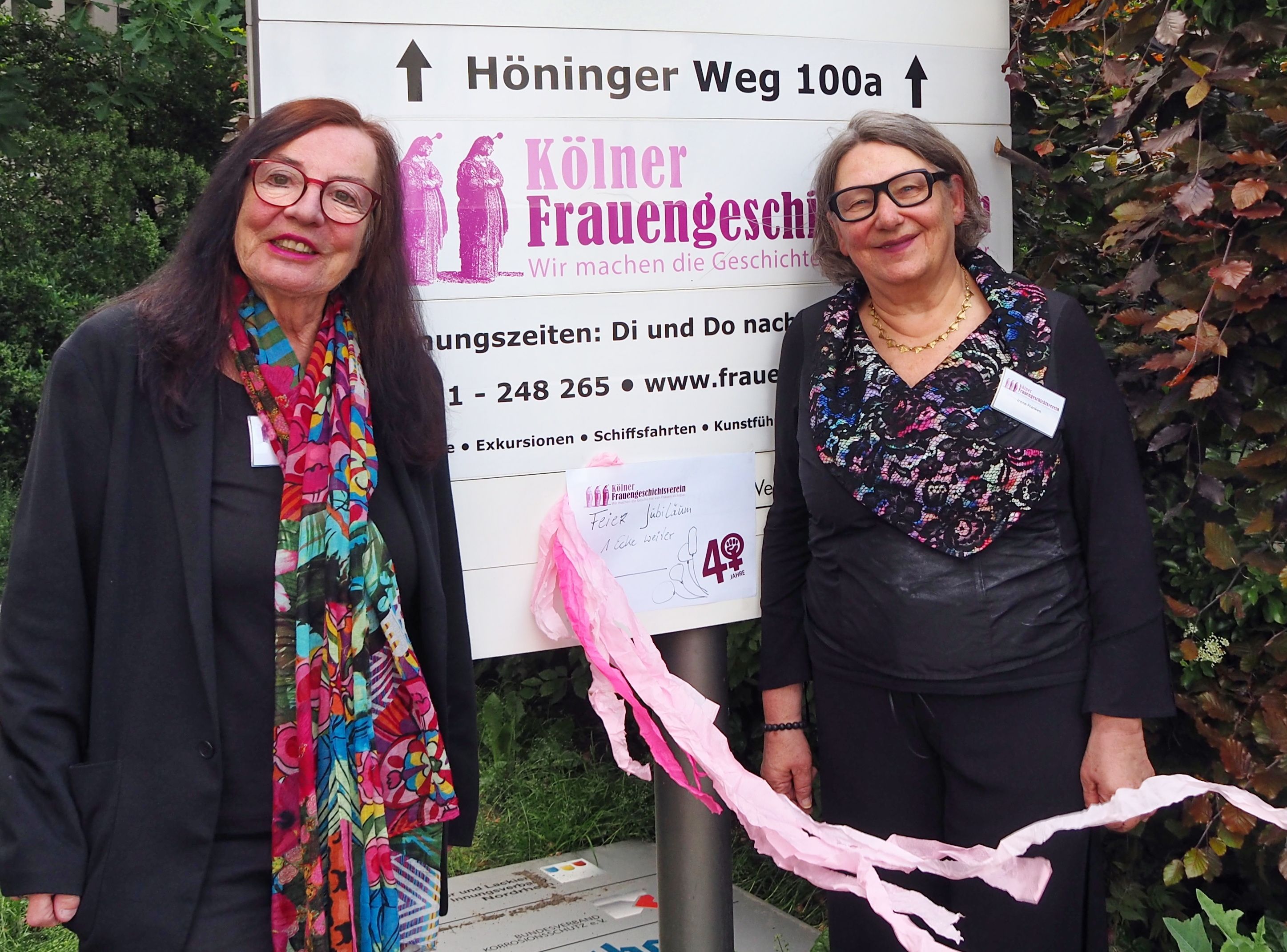 Zwei Frauen in festlicher Kleidung stehen neben dem Schild, das auf die Adresse des Kölner Frauengeschichtsvereins weist.