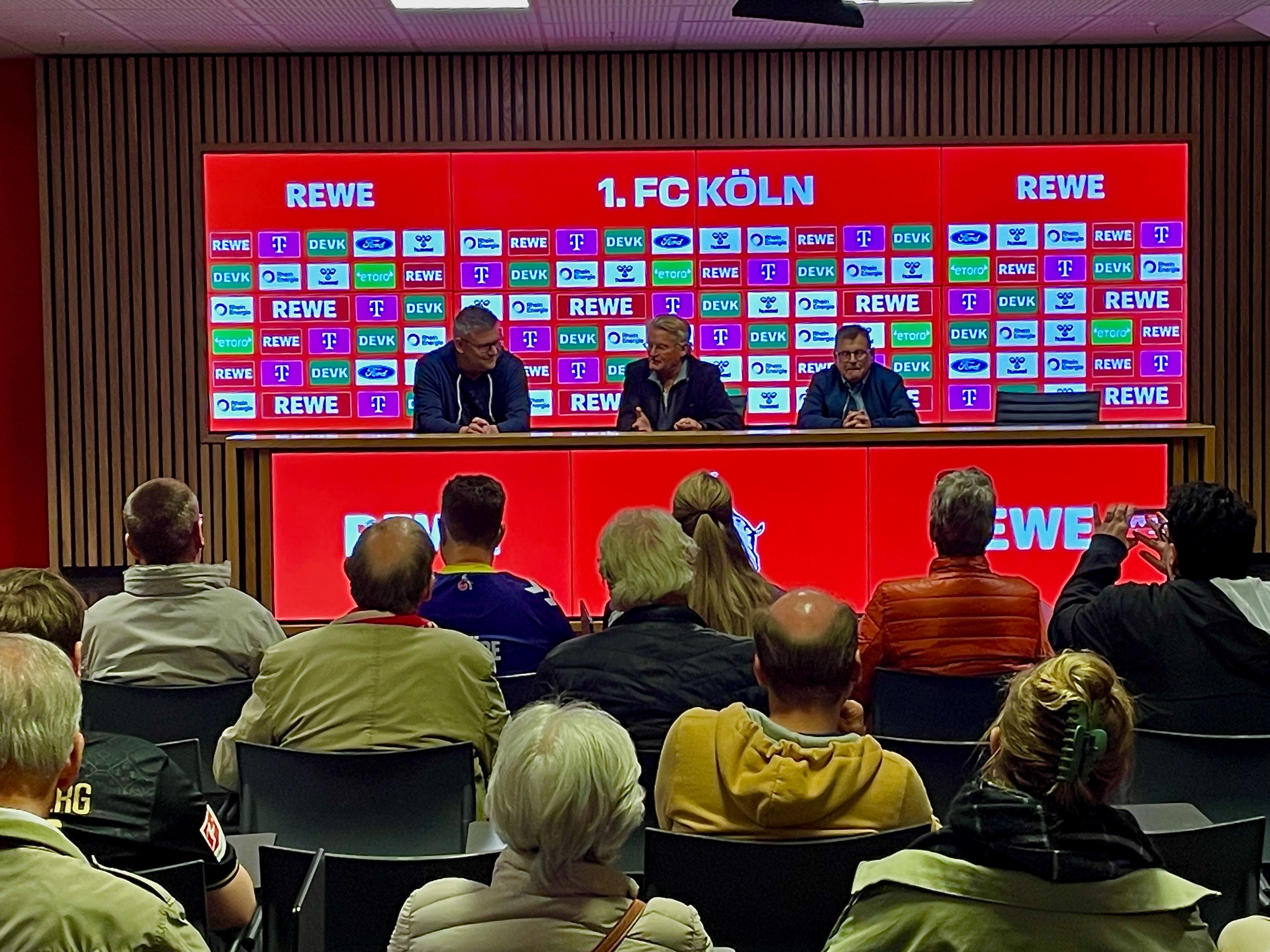 Menschen sitzen auf Stühlen und schauen auf das Set-up der FC-Pressekonferenz. Dort sitzt in der Mitte Michael Trippel.