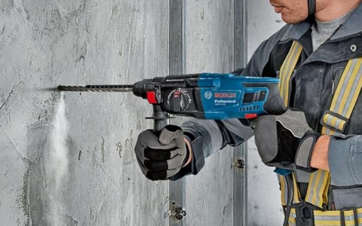 Ein Handwerker in Arbeitskleidung und Schutzhandschuhen bohrt mit dem blauen Bosch Professional Bohrhammer GBH 2-21 in eine Betonwand, aus der Staub aufwirbelt. Das Bosch Professional Logo ist gut sichtbar auf dem Gerät zu erkennen.
