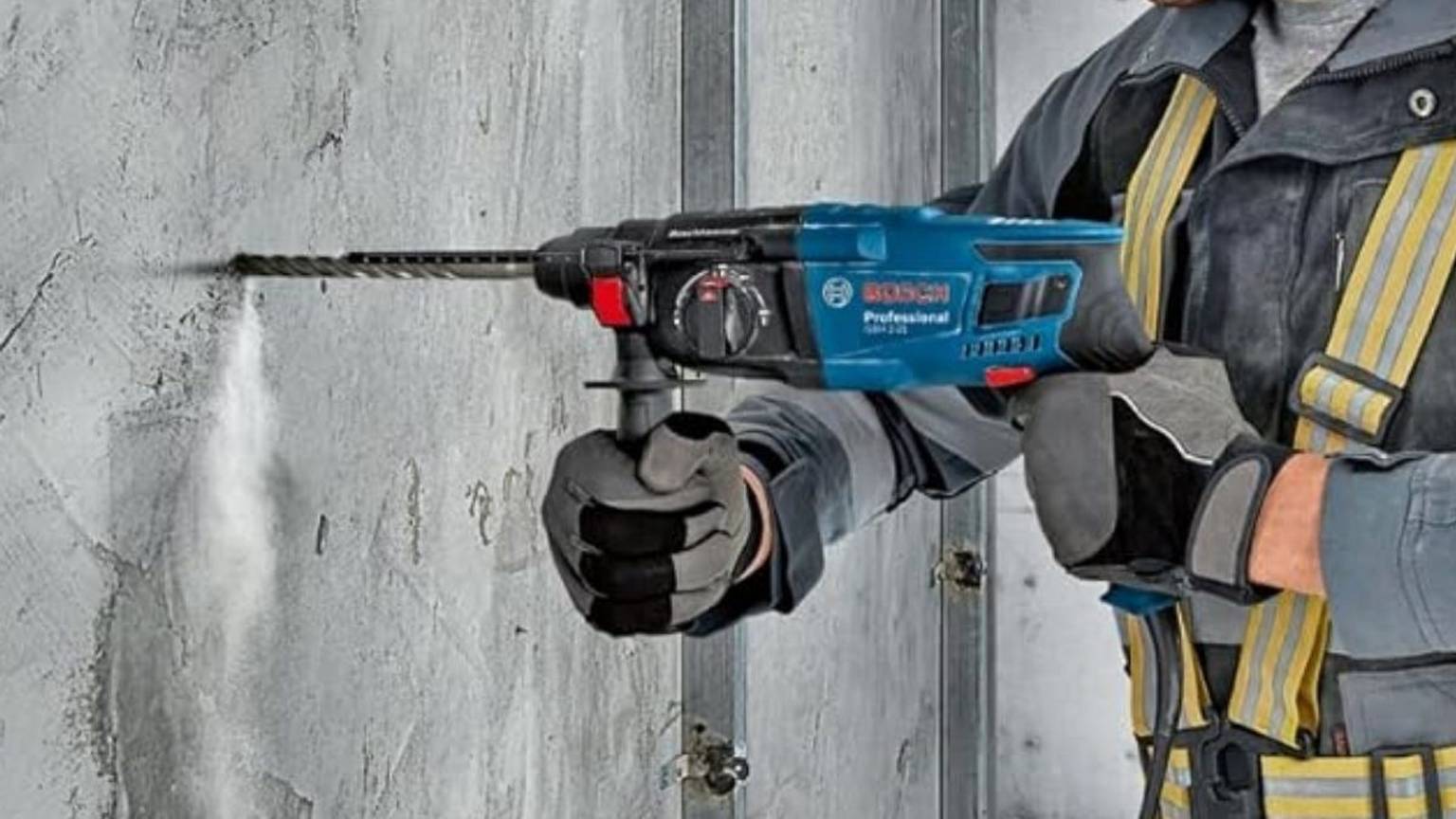 Ein Handwerker in Arbeitskleidung und Schutzhandschuhen bohrt mit dem blauen Bosch Professional Bohrhammer GBH 2-21 in eine Betonwand, aus der Staub aufwirbelt. Das Bosch Professional Logo ist gut sichtbar auf dem Gerät zu erkennen.