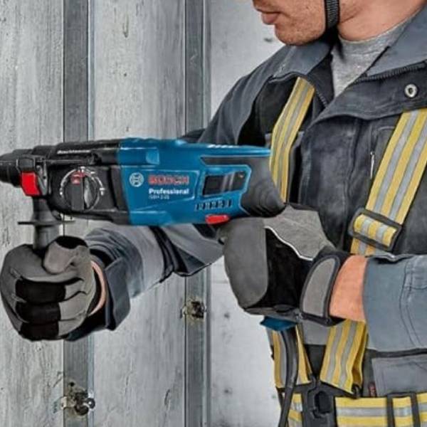 Ein Handwerker in Arbeitskleidung und Schutzhandschuhen bohrt mit dem blauen Bosch Professional Bohrhammer GBH 2-21 in eine Betonwand, aus der Staub aufwirbelt. Das Bosch Professional Logo ist gut sichtbar auf dem Gerät zu erkennen.