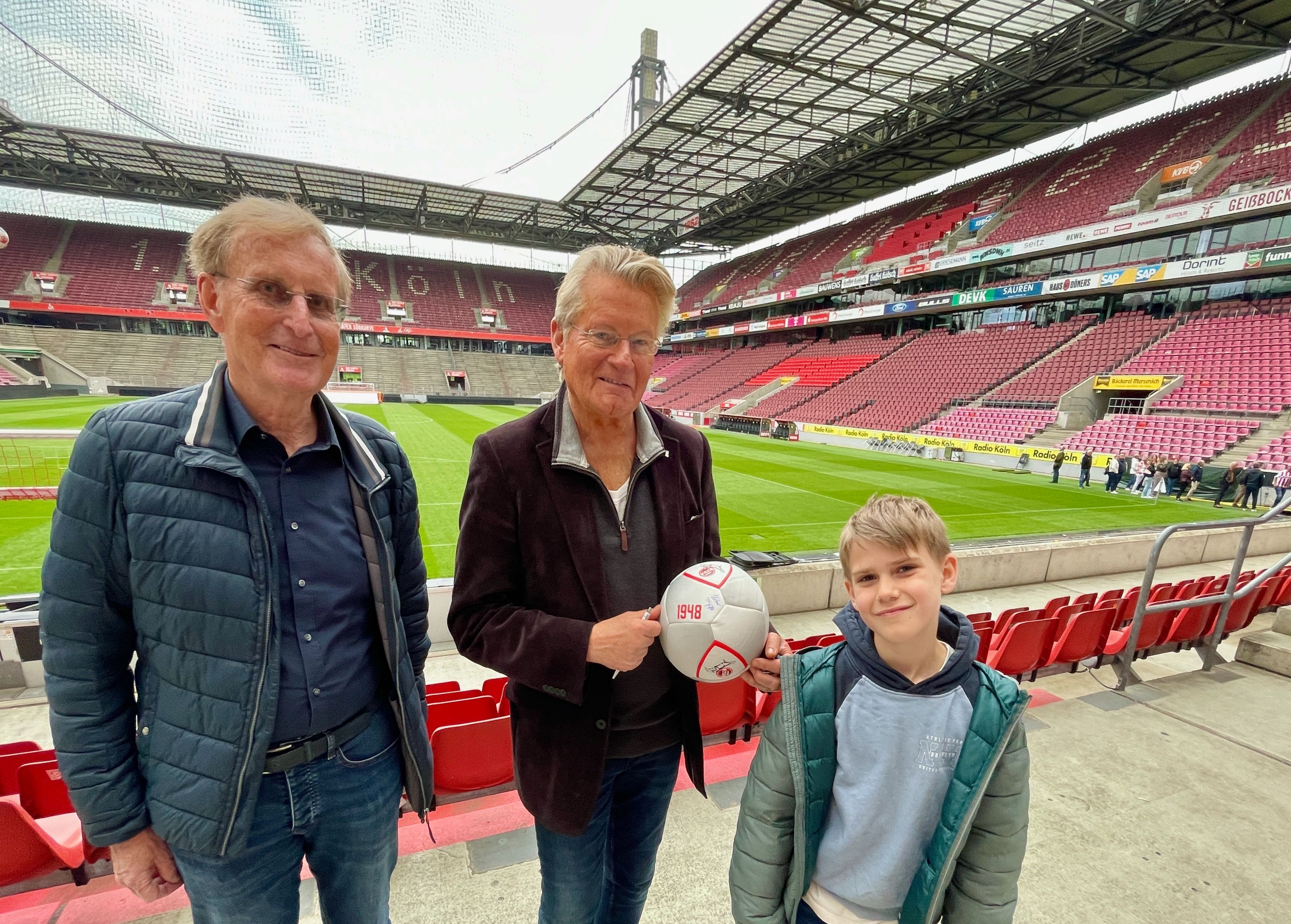 Zwei Männer und ein Junge stehen nebeneinander im Stadion. Der Mann in der Mitte hält einen Fußball.