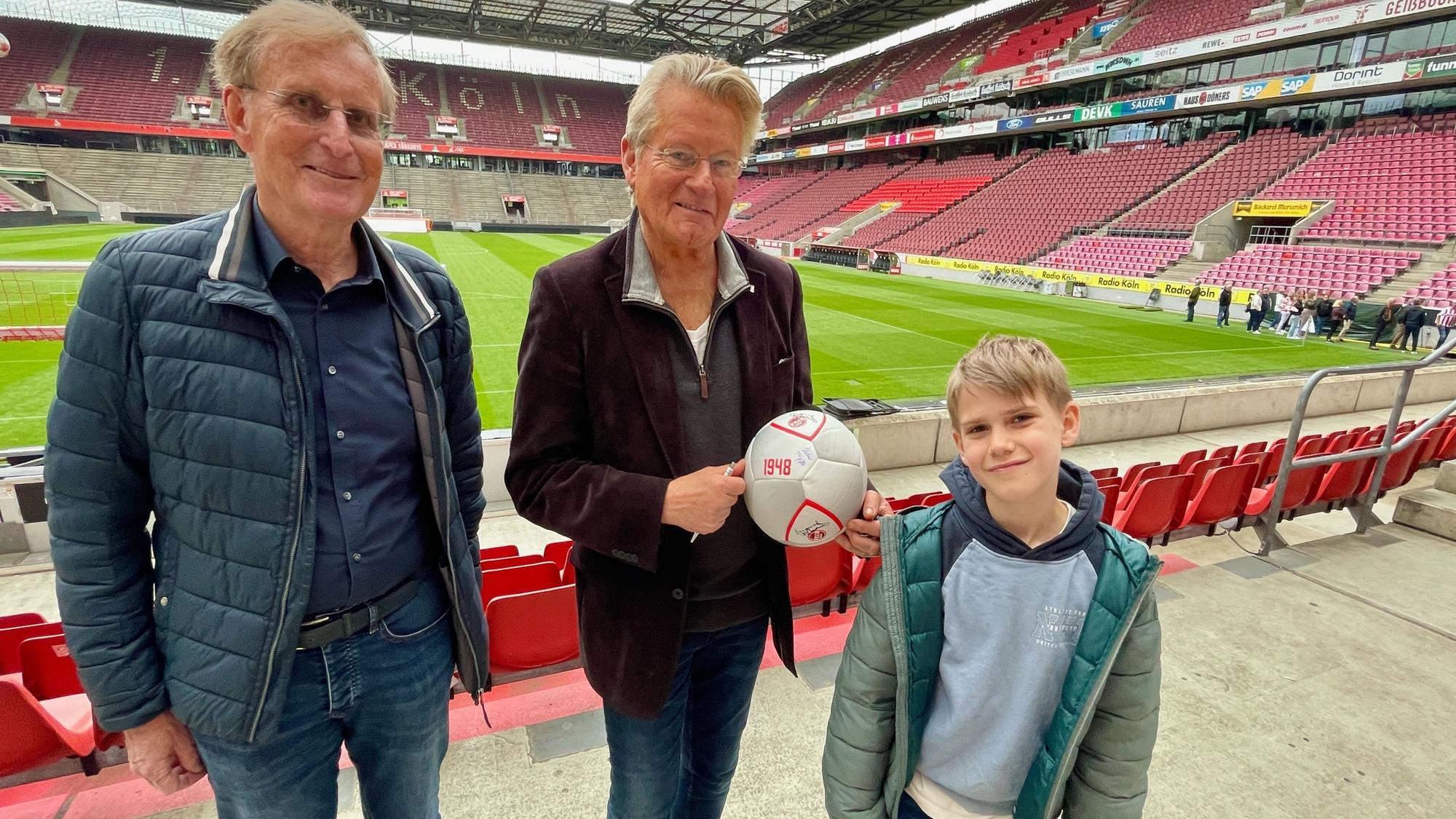 Zwei Männer und ein Junge stehen nebeneinander im Stadion. Der Mann in der Mitte hält einen Fußball.