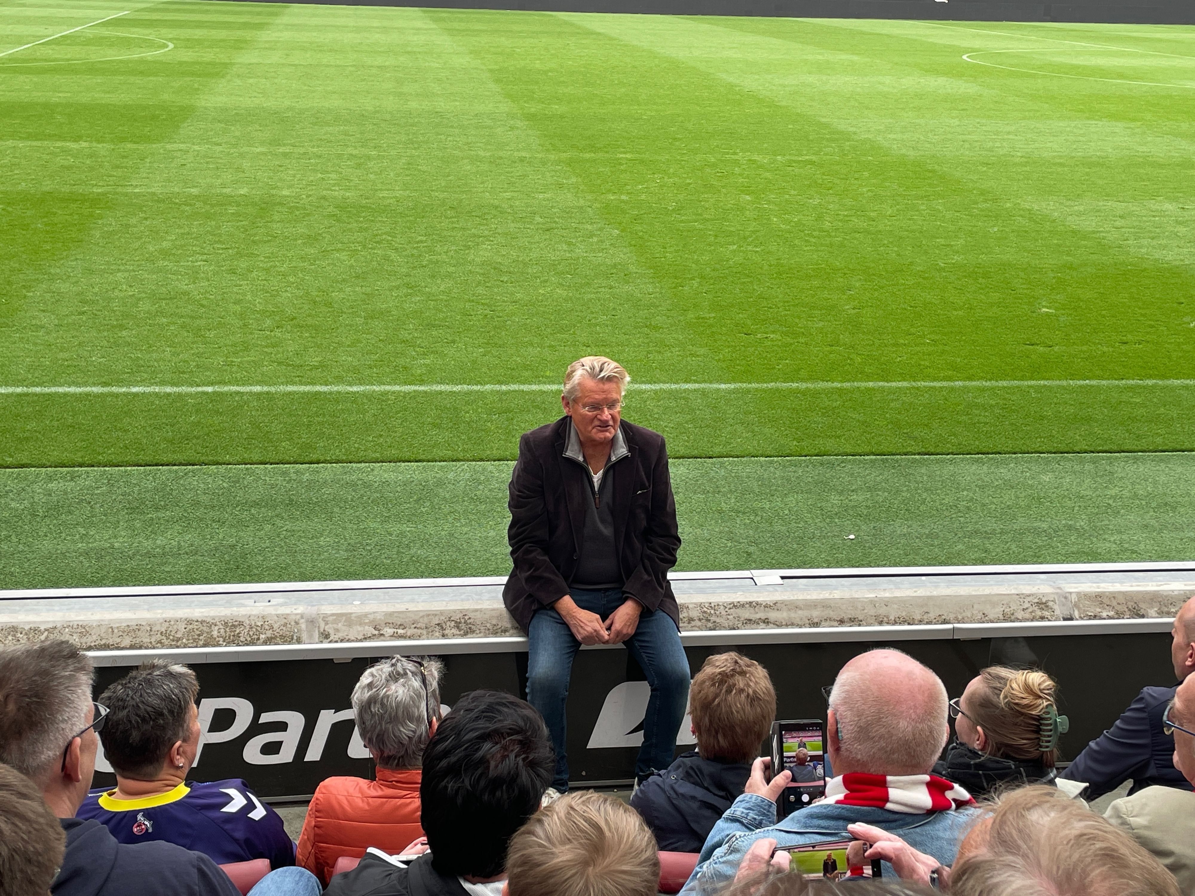 Ein Mann sitzt am Rand der Tribünenbegrenzung im Stadion und spricht mit den Menschen, die vor ihm sitzen.